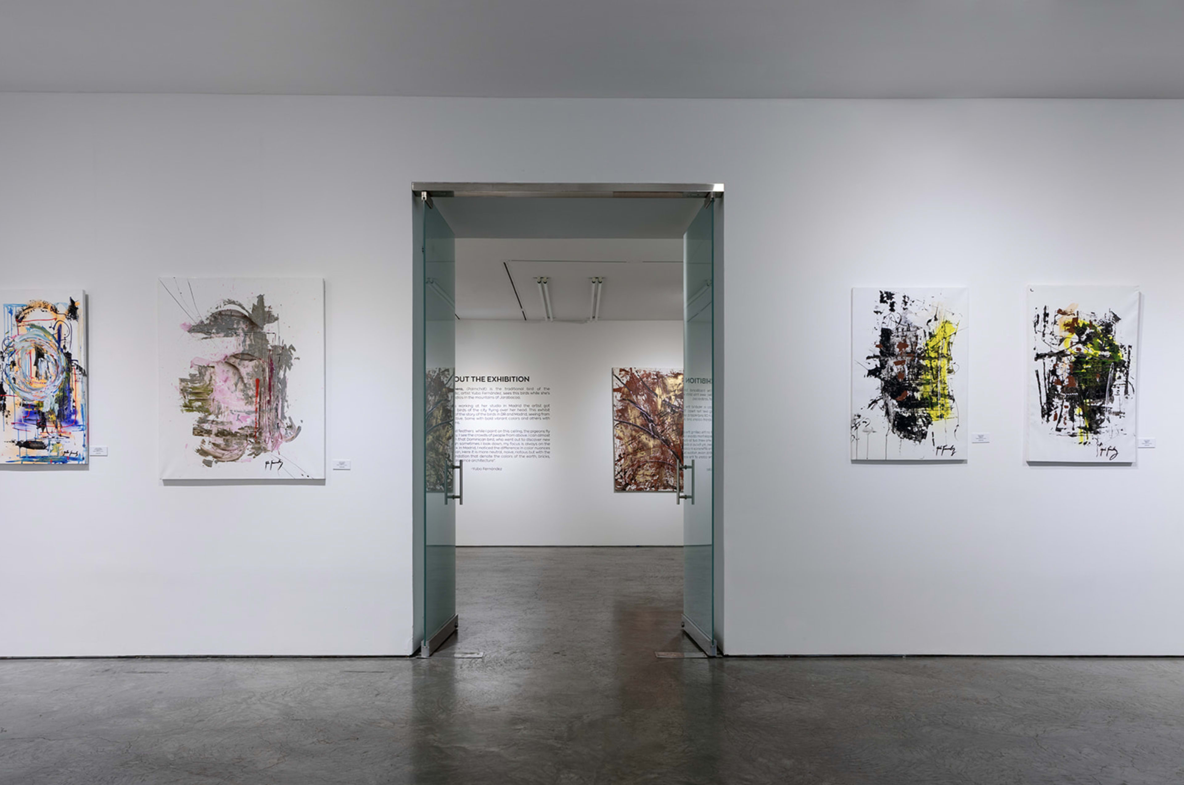 The image shows a modern art gallery with white walls displaying several abstract paintings on either side of an entrance that leads to a text panel and additional artworks in the background.