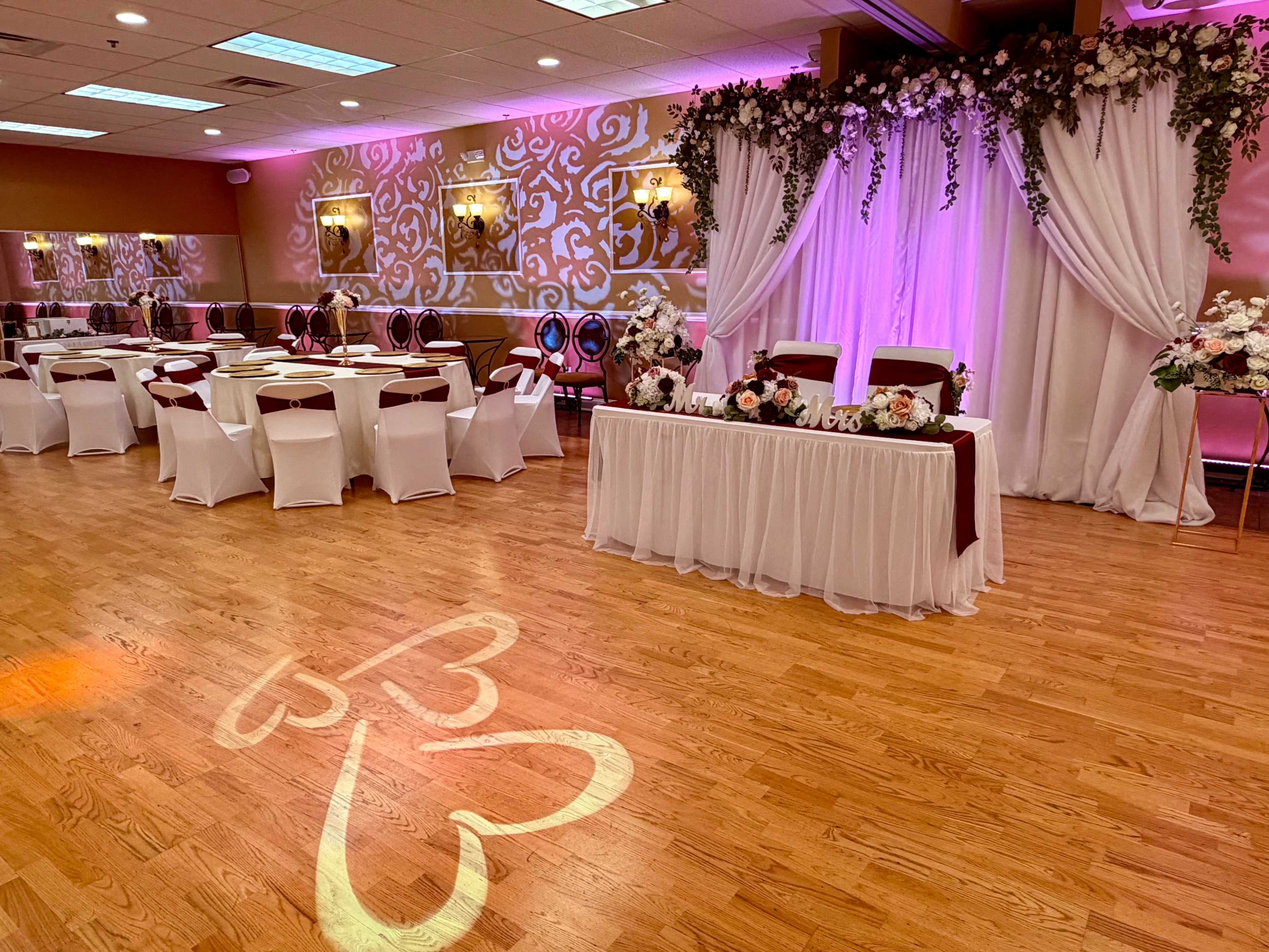 The image shows a banquet hall set up for an event, featuring elegantly arranged tables with white chairs, a decorated stage area with floral accents, and soft pink lighting.