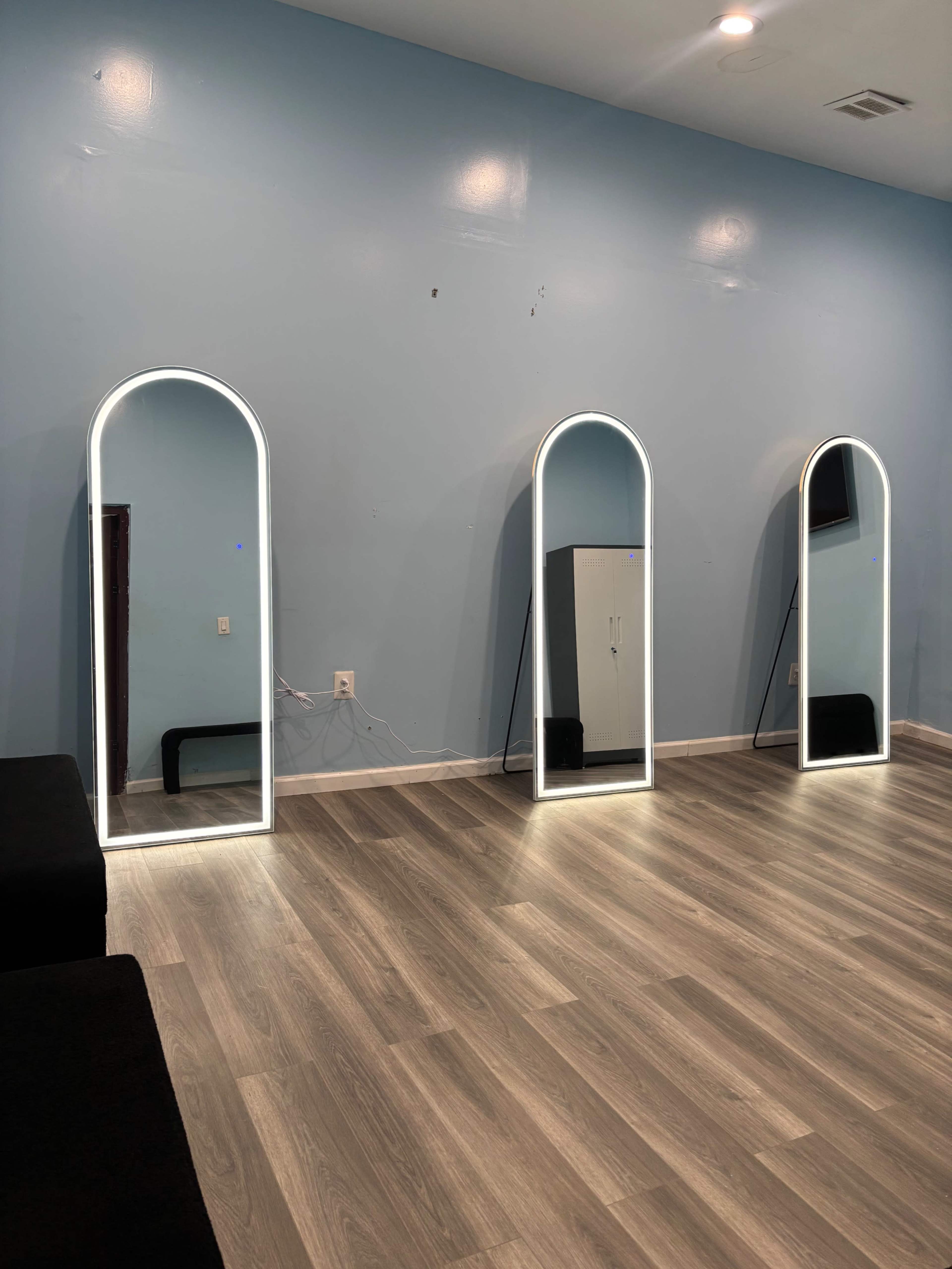 The image shows three arched mirrors with LED lighting against a light blue wall in a room with wooden flooring.