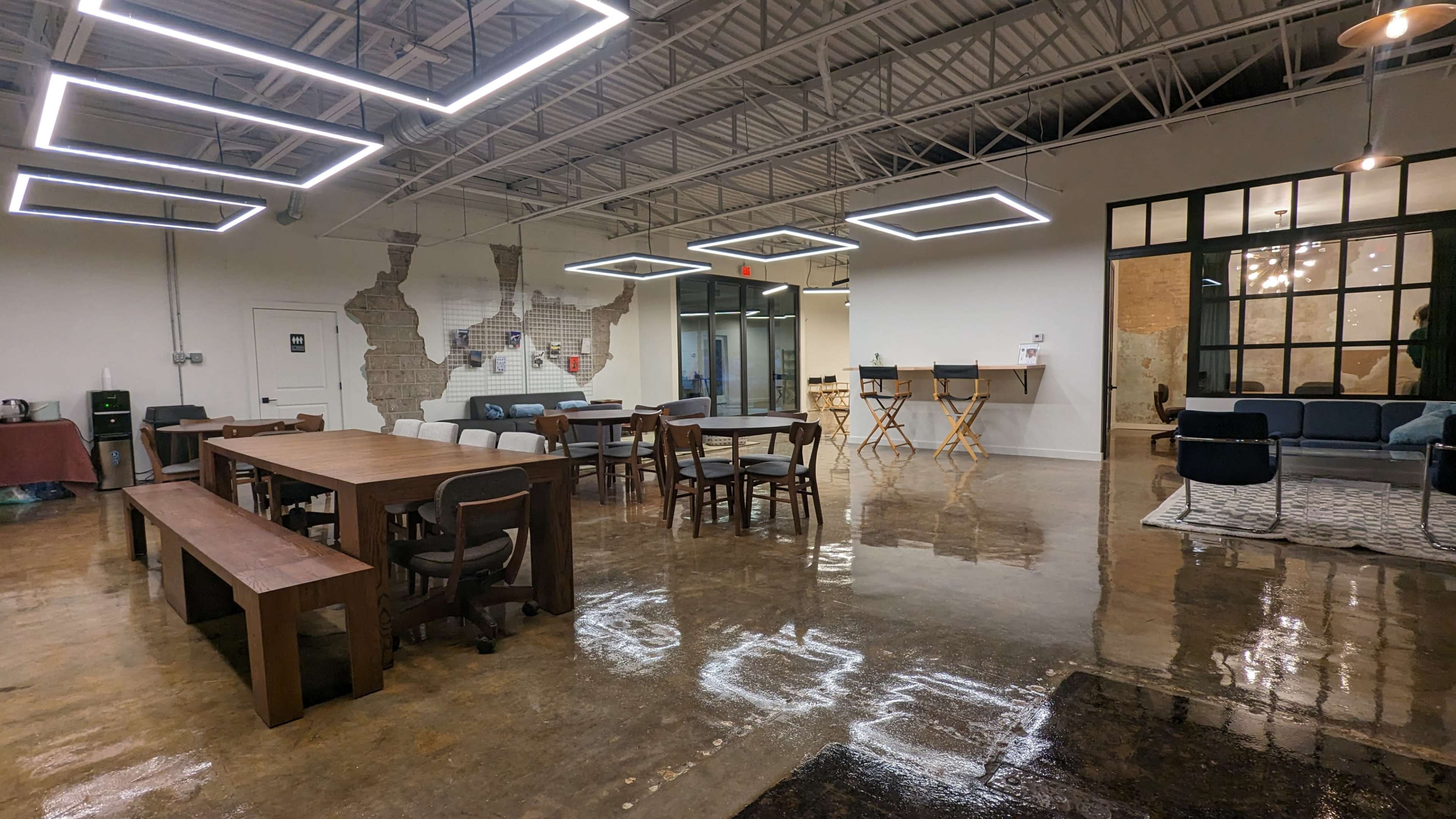 The image shows a modern, open workspace featuring a mix of wooden furniture, including dining tables and chairs, with polished concrete floors and stylish lighting fixtures.