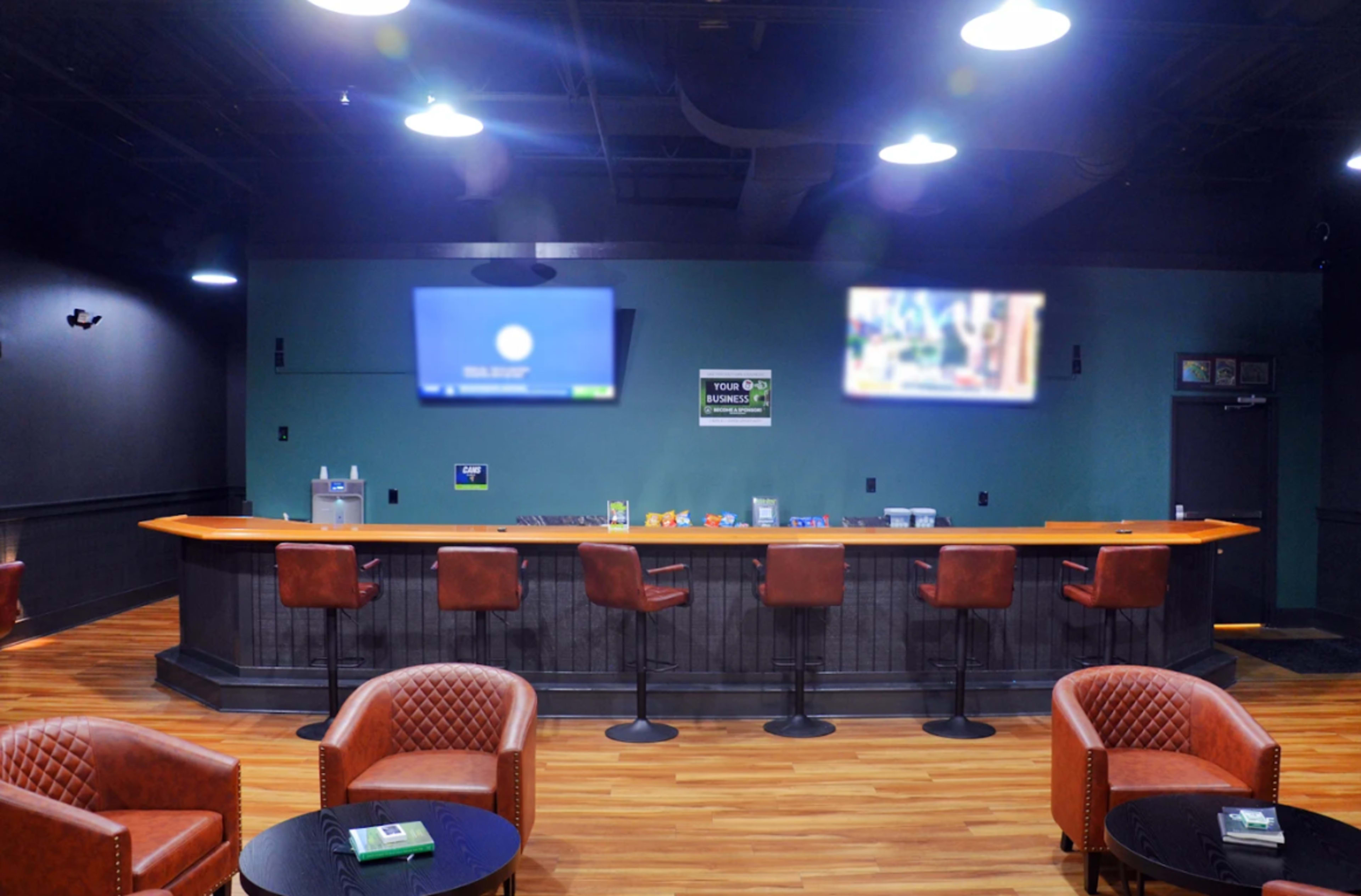 The image shows a modern bar area featuring two flat-screen TVs mounted on the wall, a long orange bar counter, and several brown cushioned chairs arranged around small black tables.