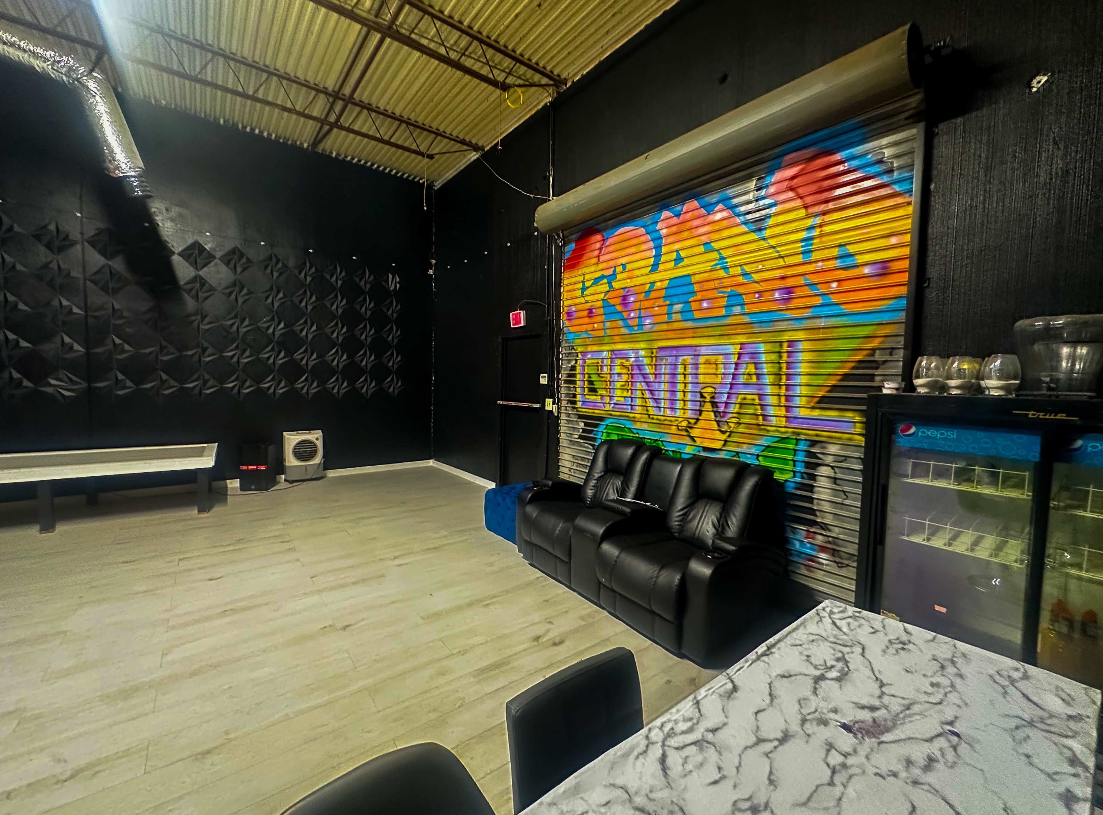 The image shows a dimly lit room with black walls, featuring a colorful mural, a black couch, a table with a marble top, and a small fridge against one wall.