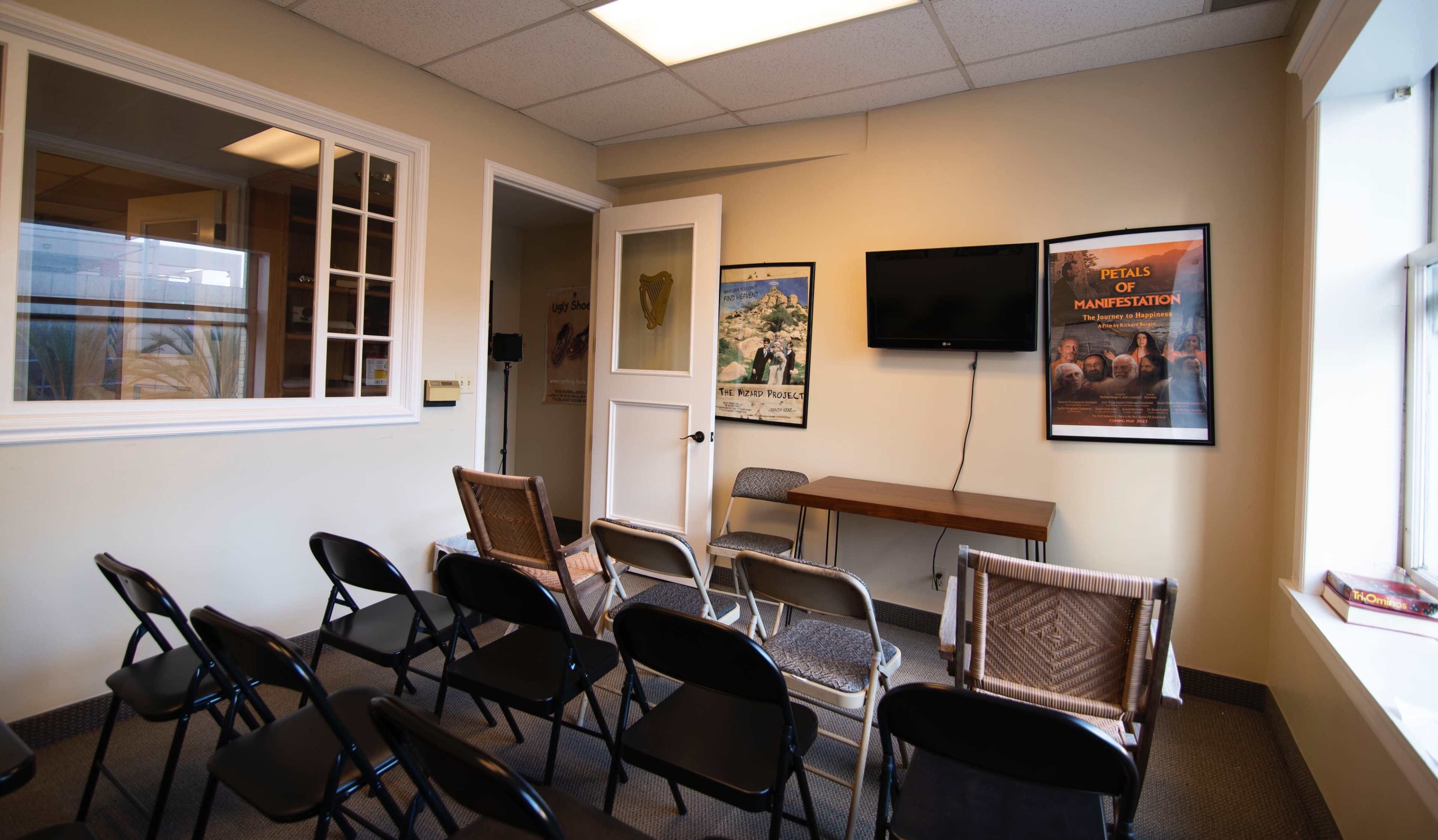 A small meeting room with rows of folding chairs facing a wall-mounted television and a table against the opposite wall, adorned with posters.