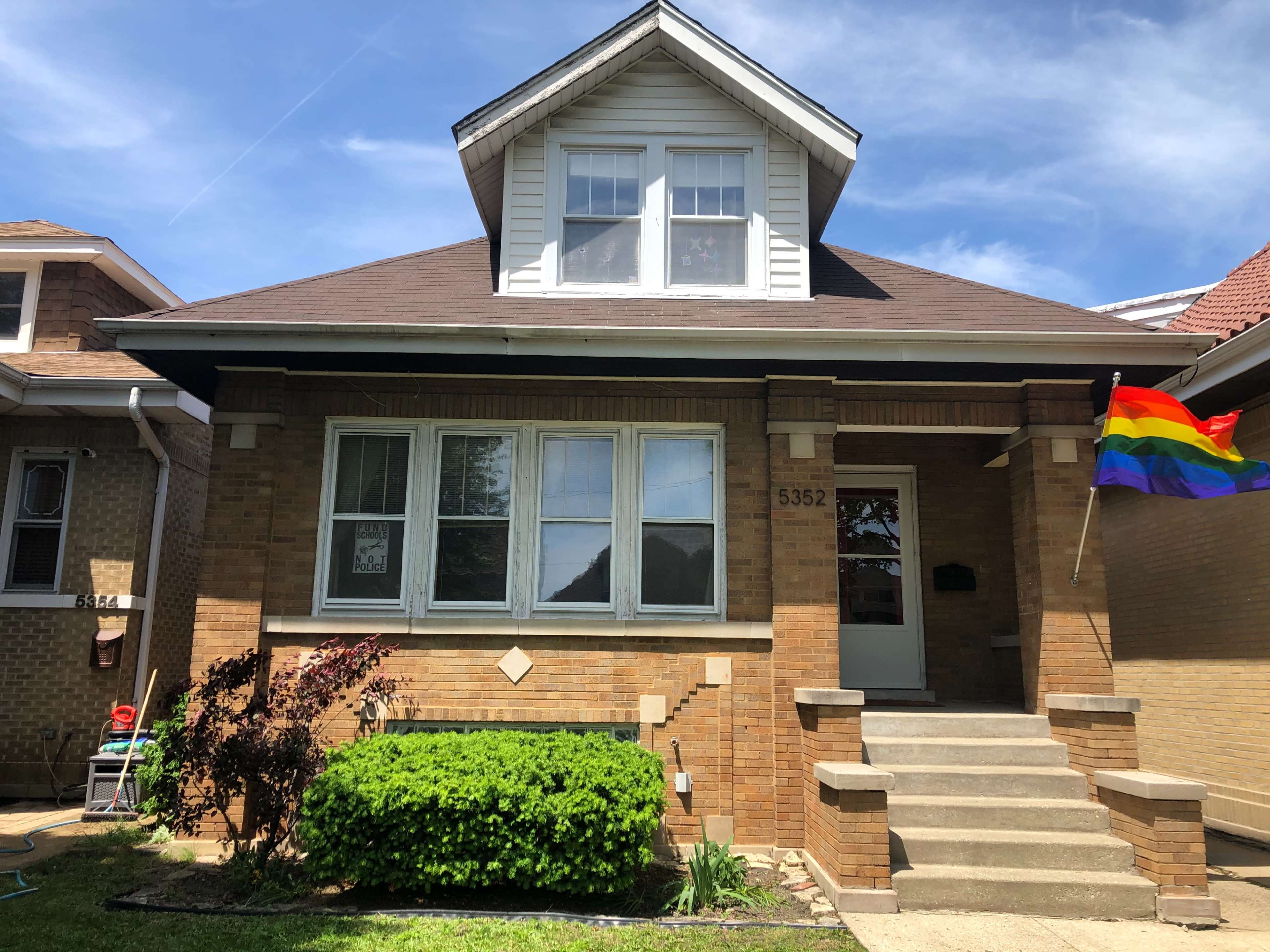 A two-story brick house with a front porch displays a rainbow flag next to well-maintained shrubs and lawn.