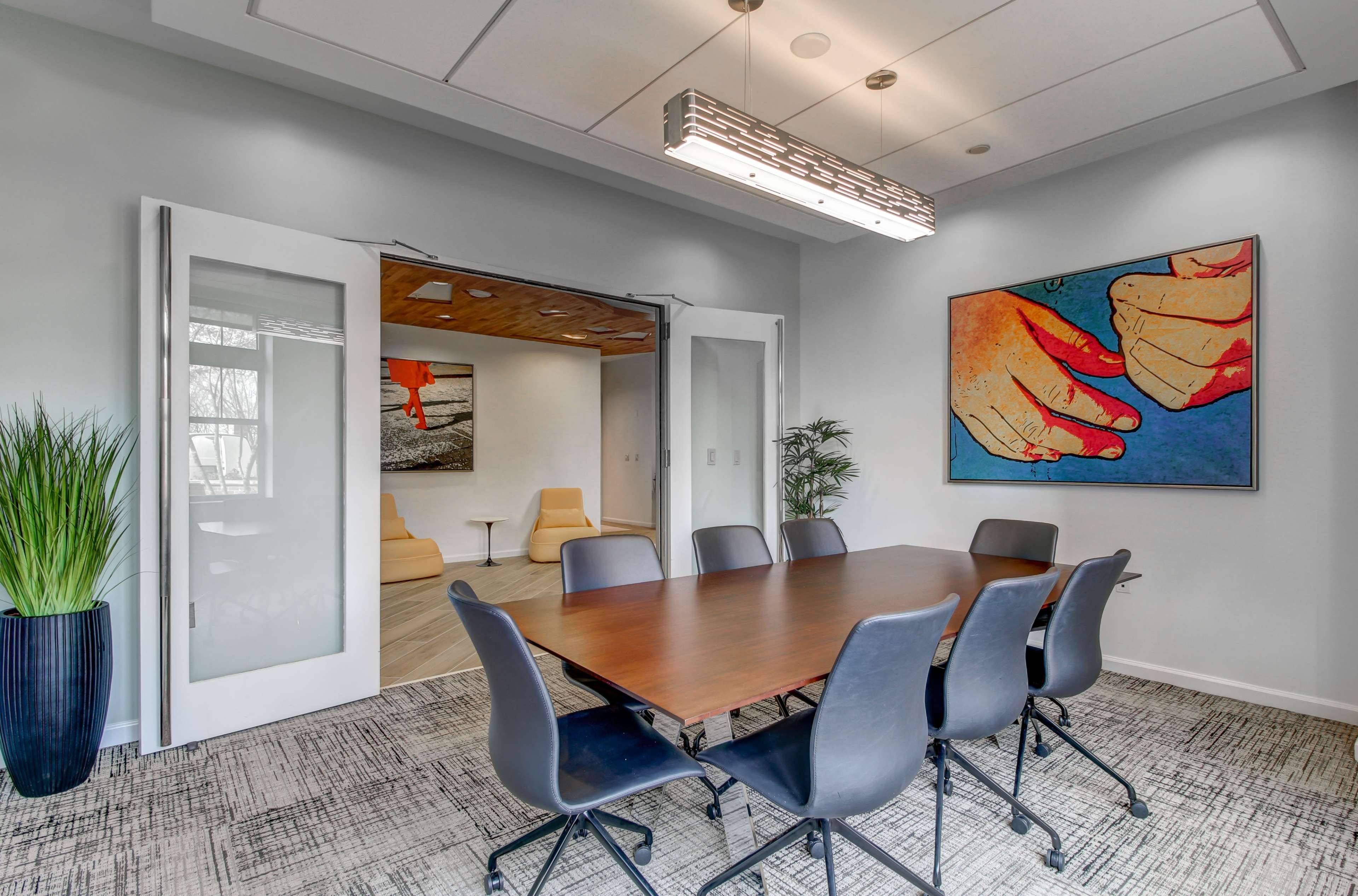 A conference room features a large wooden table surrounded by black chairs, with a contemporary artwork on the wall and a view into a lounge area through open double doors.