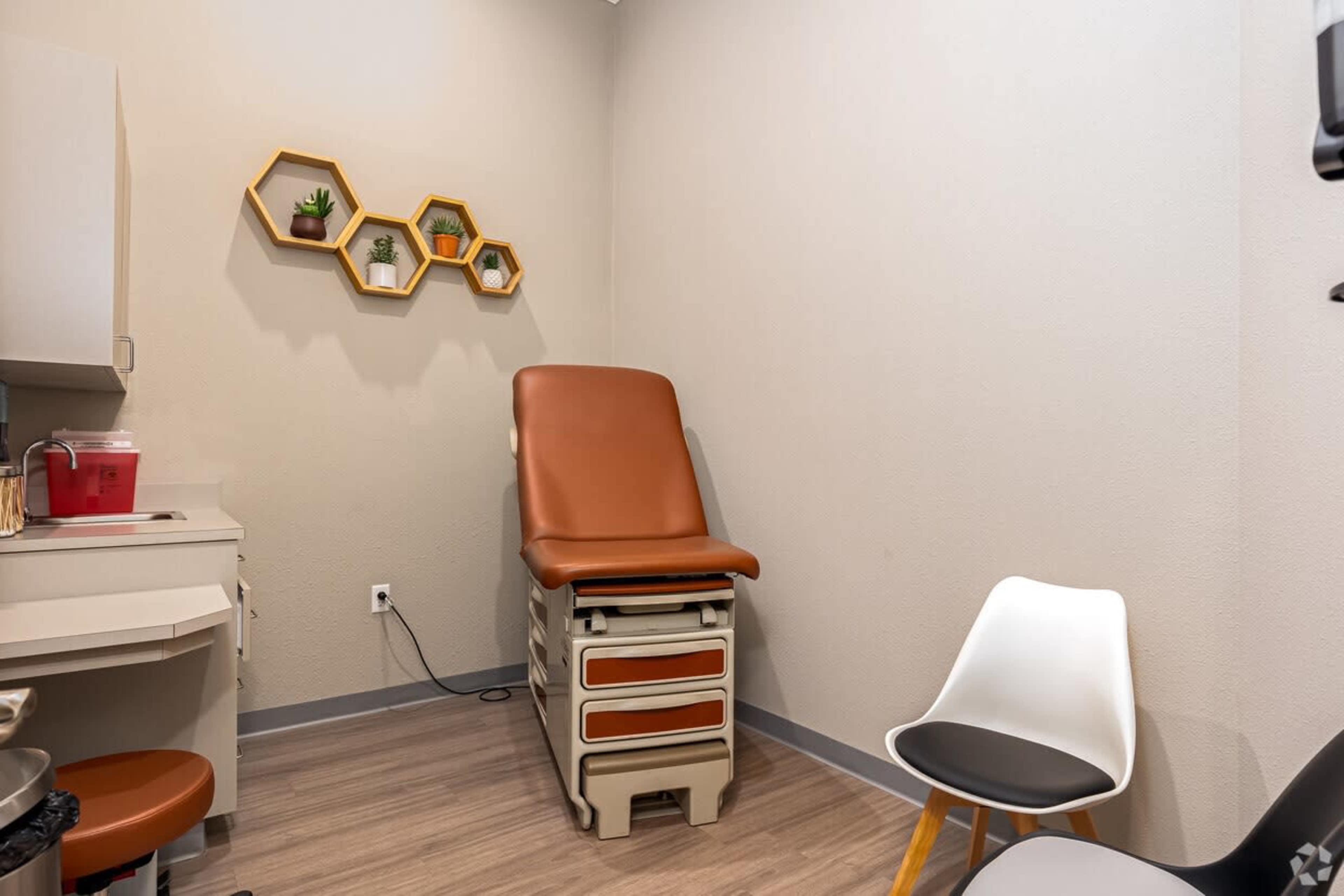 The image shows a small medical examination room with a brown examination table, a white chair, and a hexagonal shelf on the wall displaying small plants.