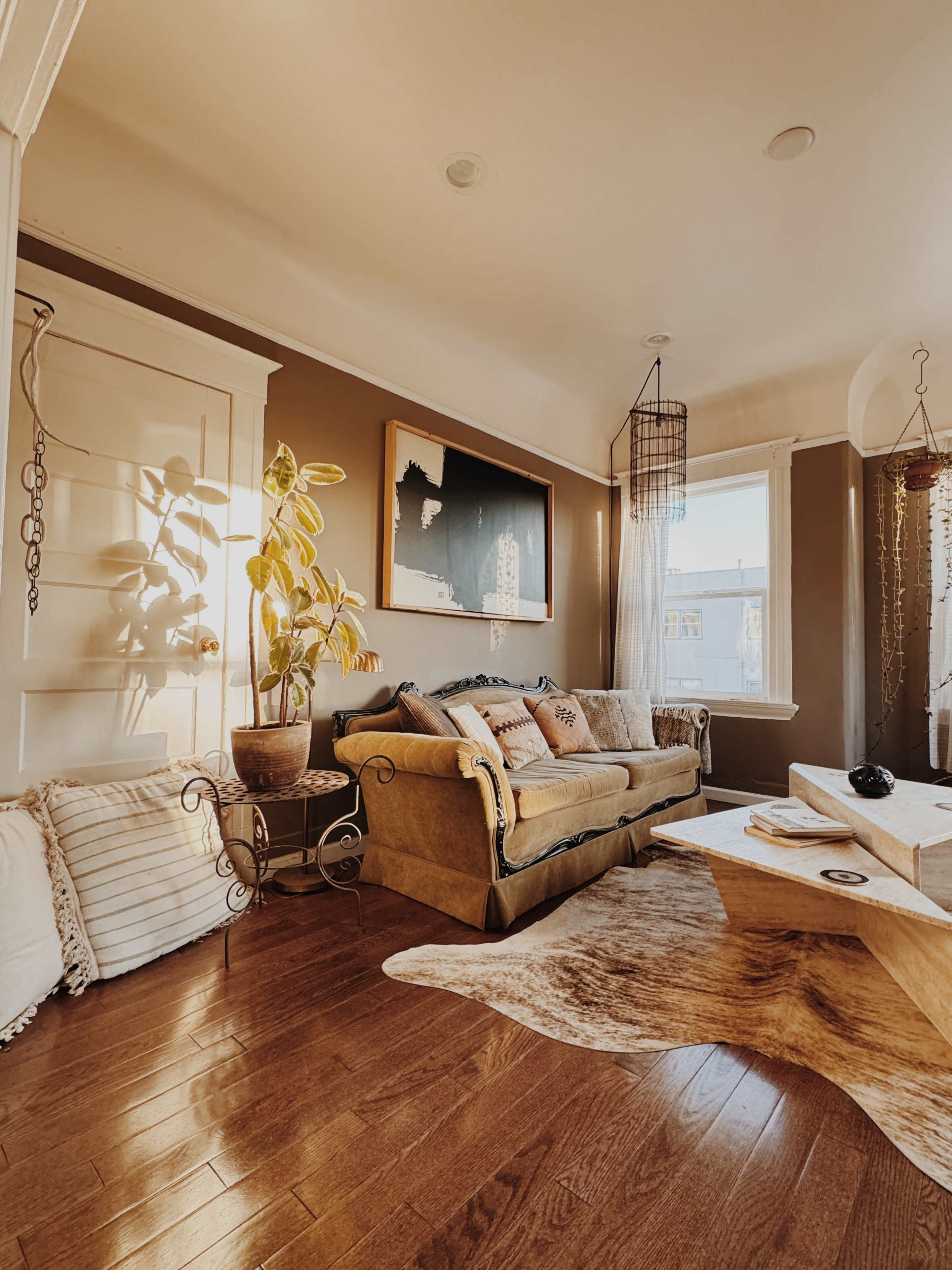The image shows a cozy living room with a sofa, decorative pillows, a coffee table, and large windows allowing natural light to brighten the space.