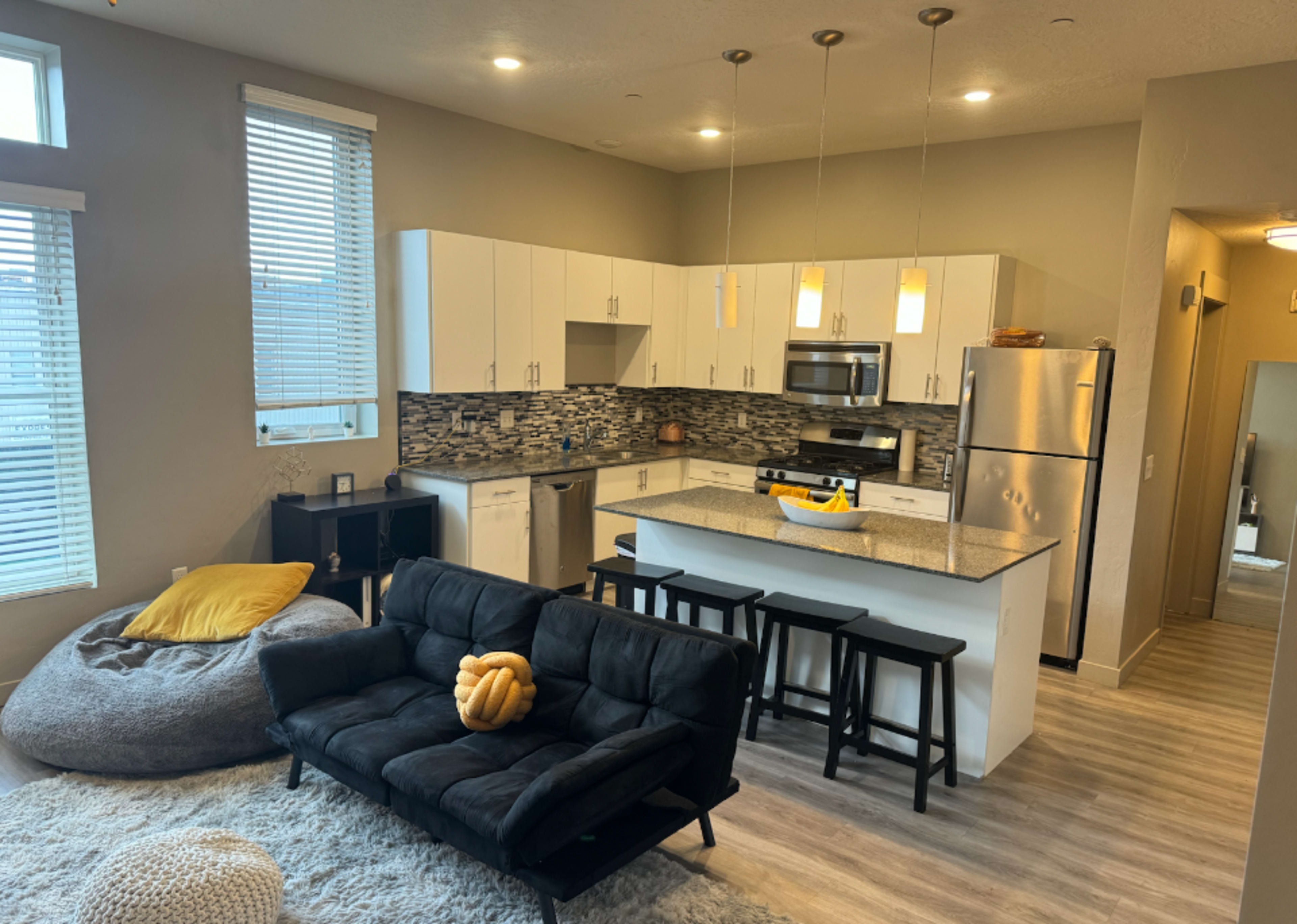 A modern open-concept kitchen and living area with white cabinets, a stainless steel refrigerator, a dark sofa, and a large bean bag.