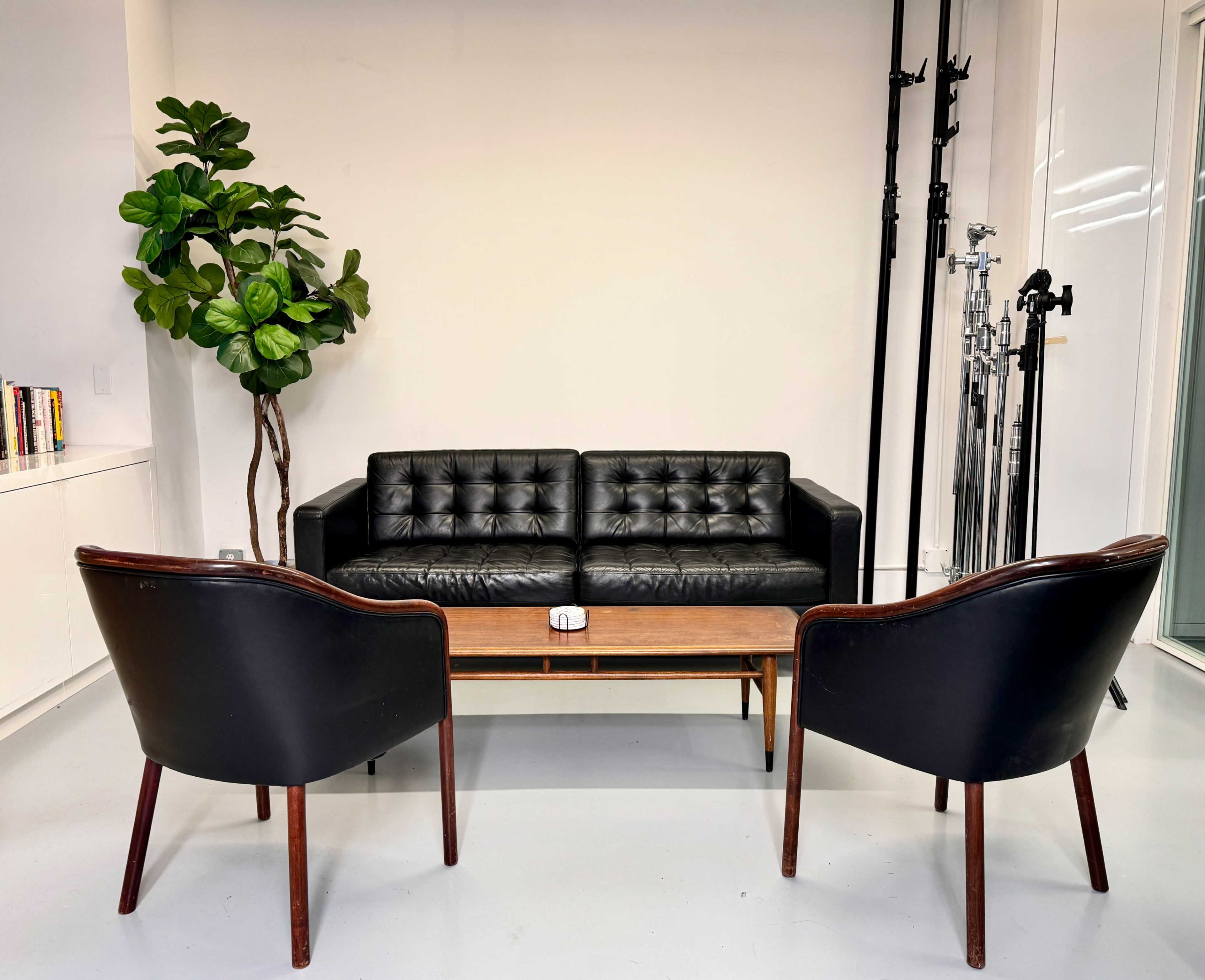 A black tufted sofa flanked by two wooden chairs, facing a coffee table in a minimalist interior setting.
