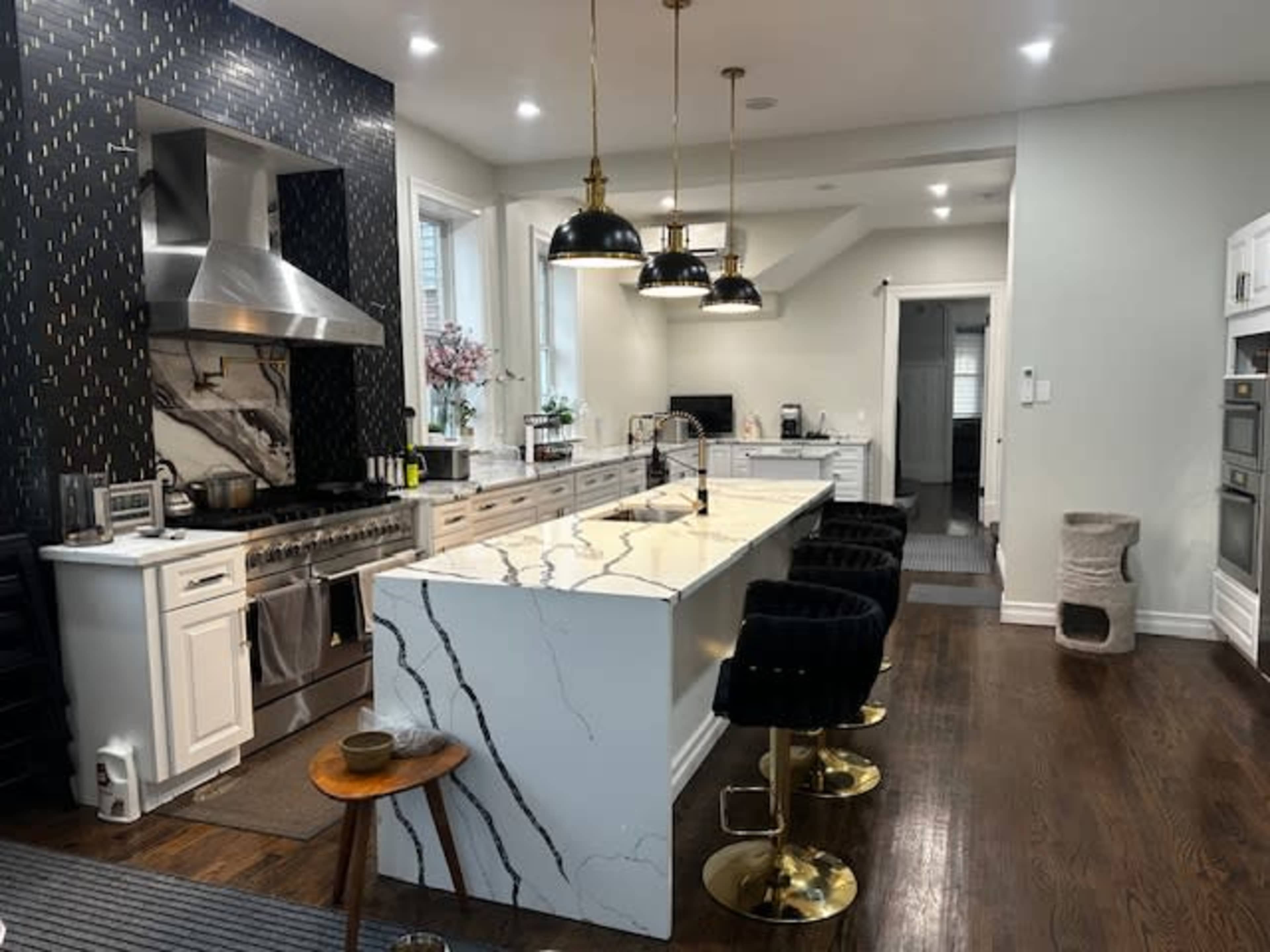 The image shows a modern kitchen featuring a large marble island with high stools, a stainless steel range hood, and black pendant lights above the counter.
