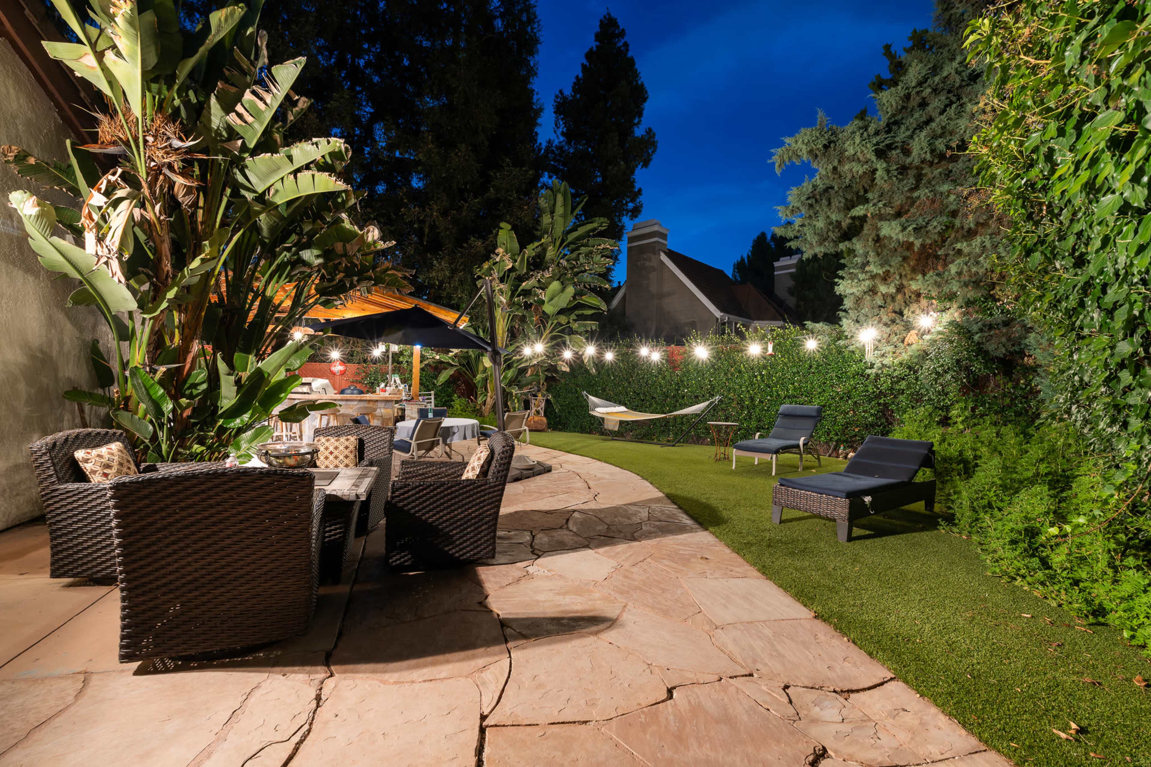 The image shows a landscaped backyard featuring a stone patio with seating, a hammock, lounge chairs, and surrounding greenery under evening lighting.
