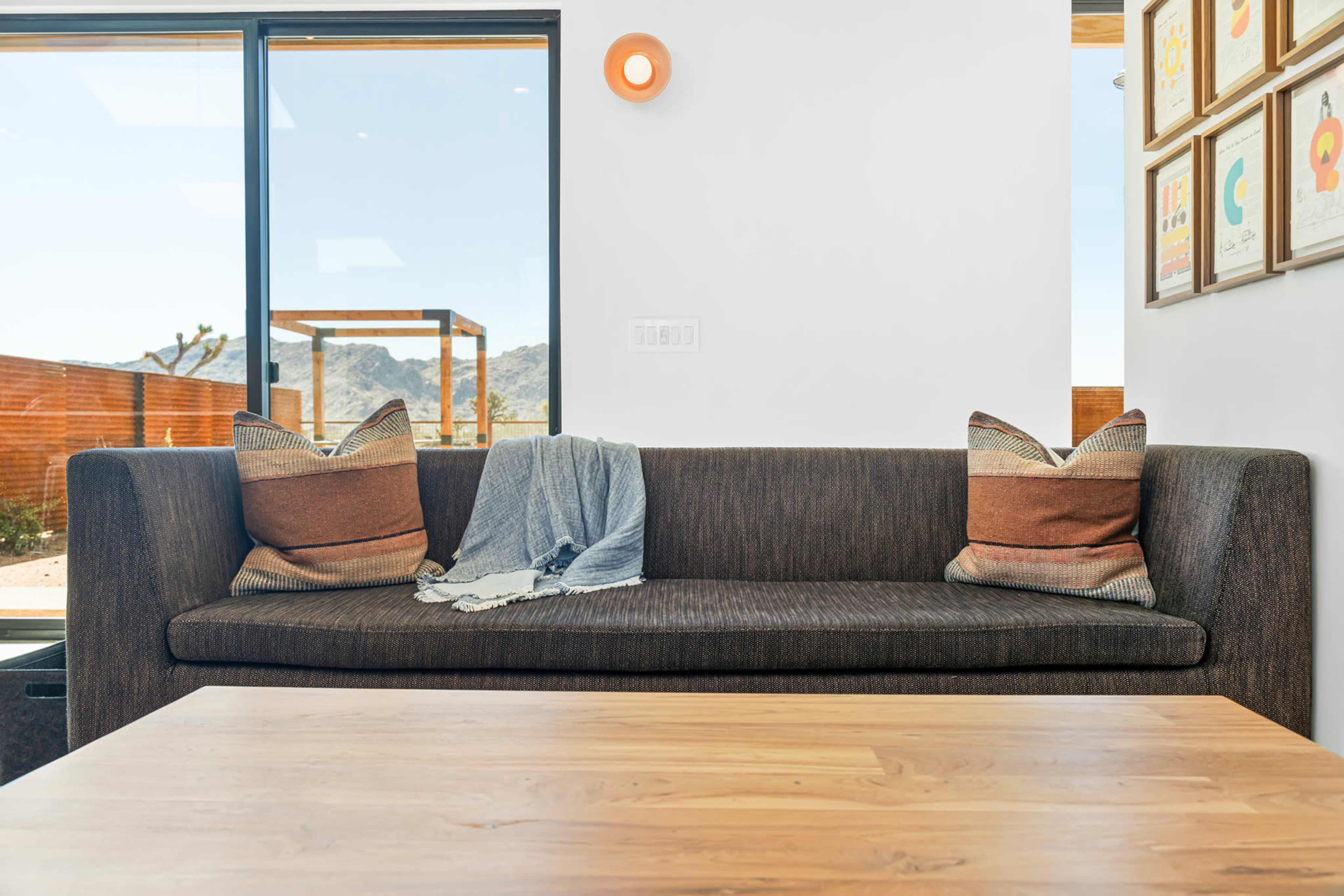 A dark sofa with decorative pillows and a draped blanket sits across from a wooden coffee table in a bright room with large windows and mountain views.