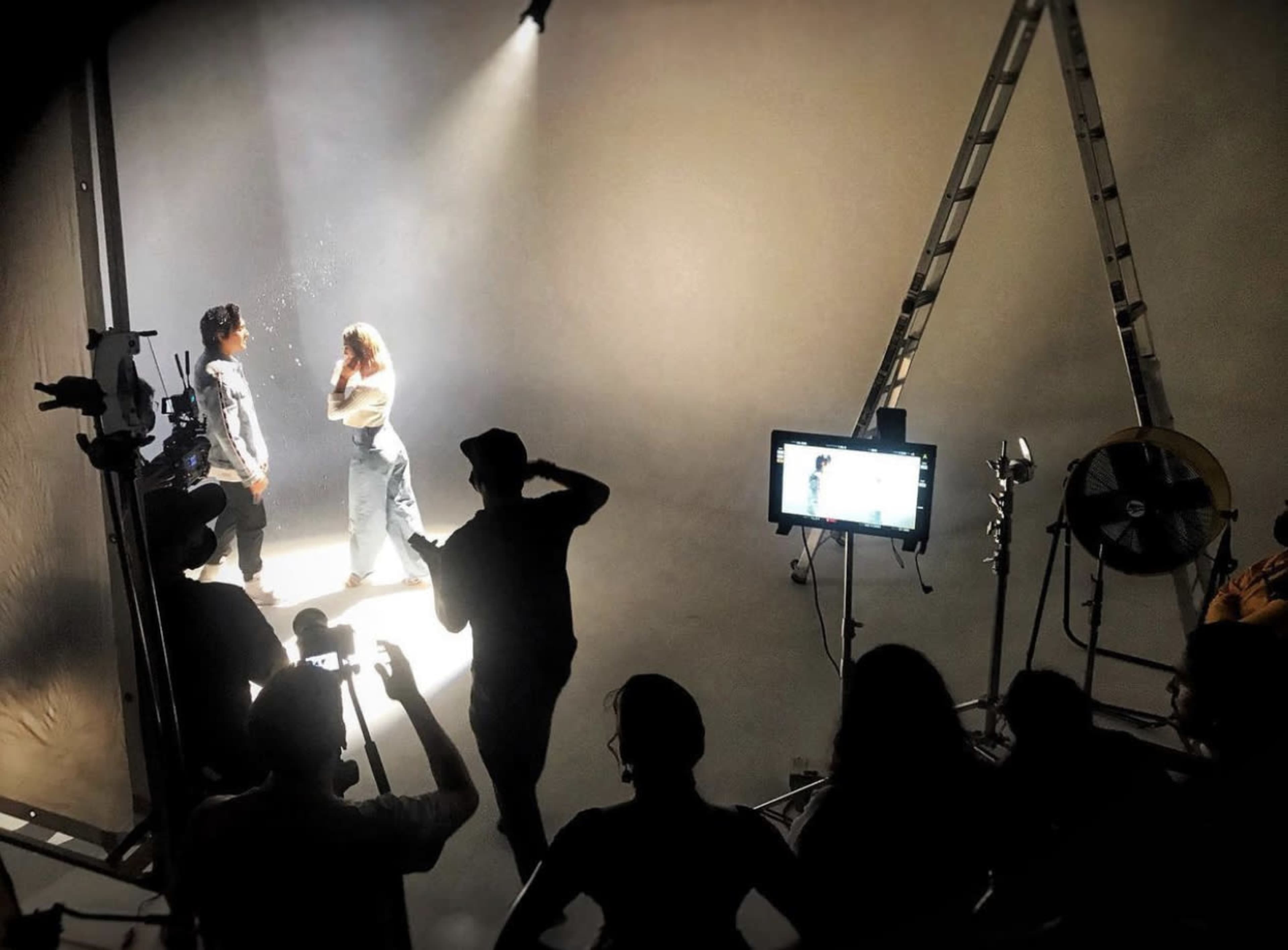 A group of people observes the filming of a scene featuring two models illuminated by soft lighting in a studio setting.