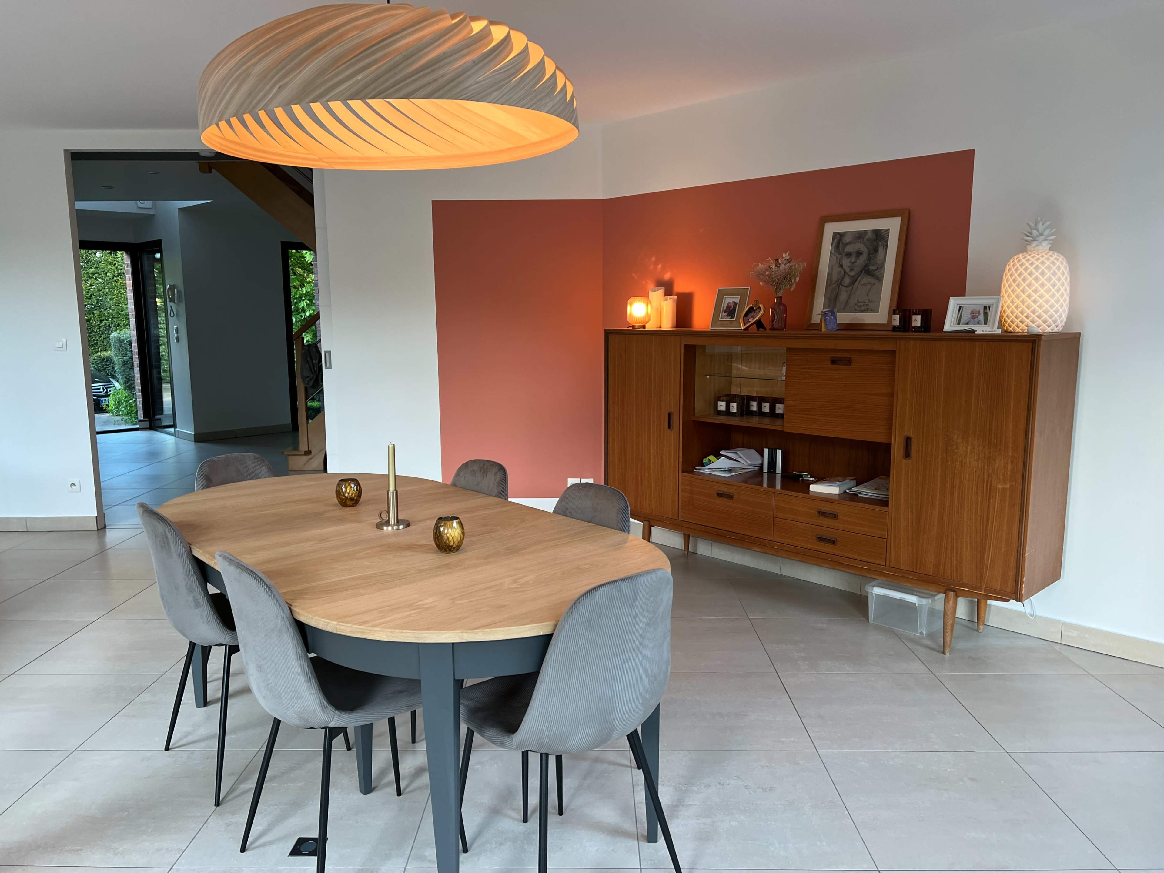 A modern dining area features a round wooden table surrounded by six gray upholstered chairs, illuminated by a large pendant light, with a side cabinet and framed artwork along the wall.