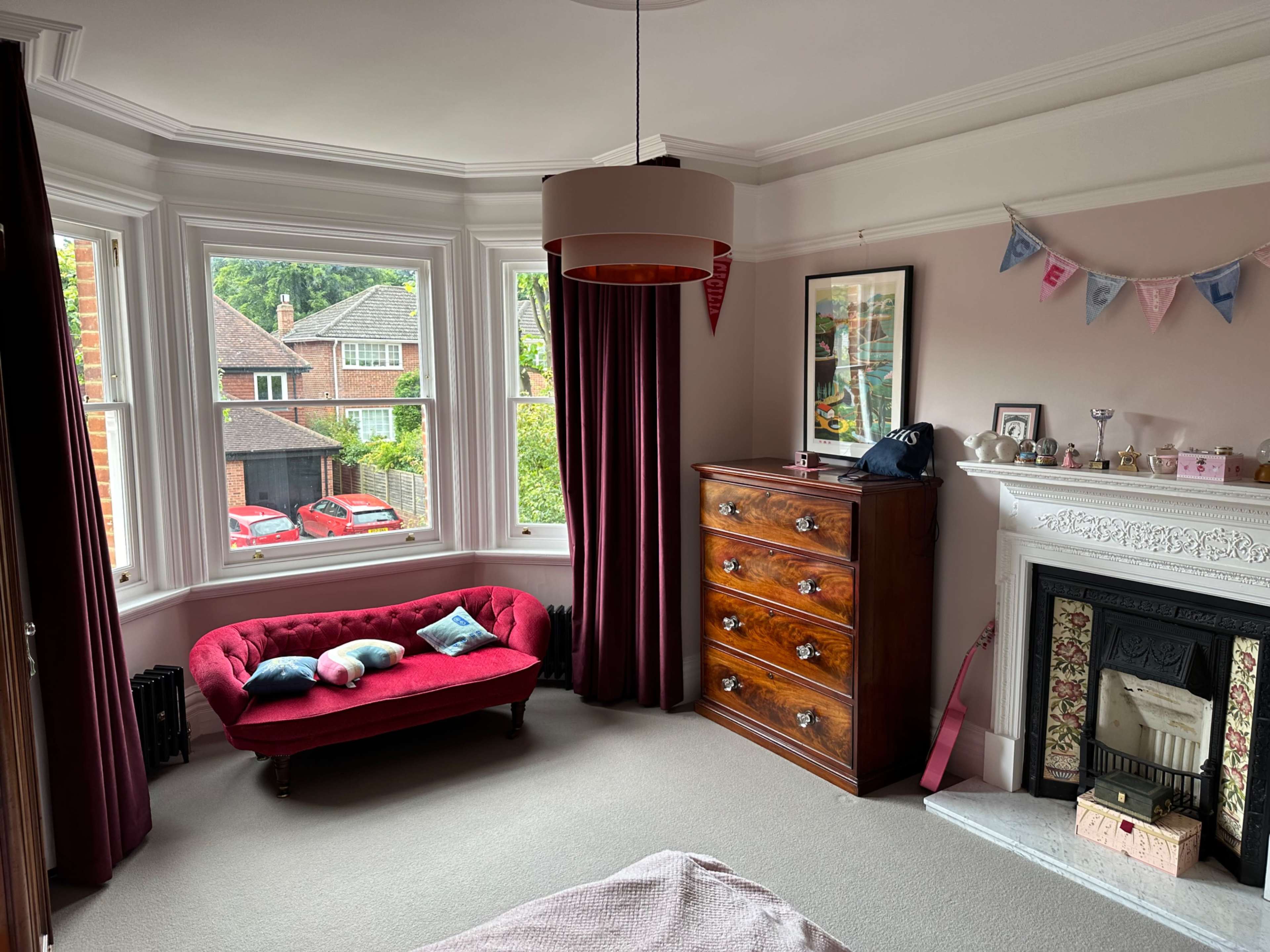 The image shows a bedroom with a red upholstered couch, a wooden dresser, and a decorative fireplace, featuring a large window with curtains and a view of the outdoors.