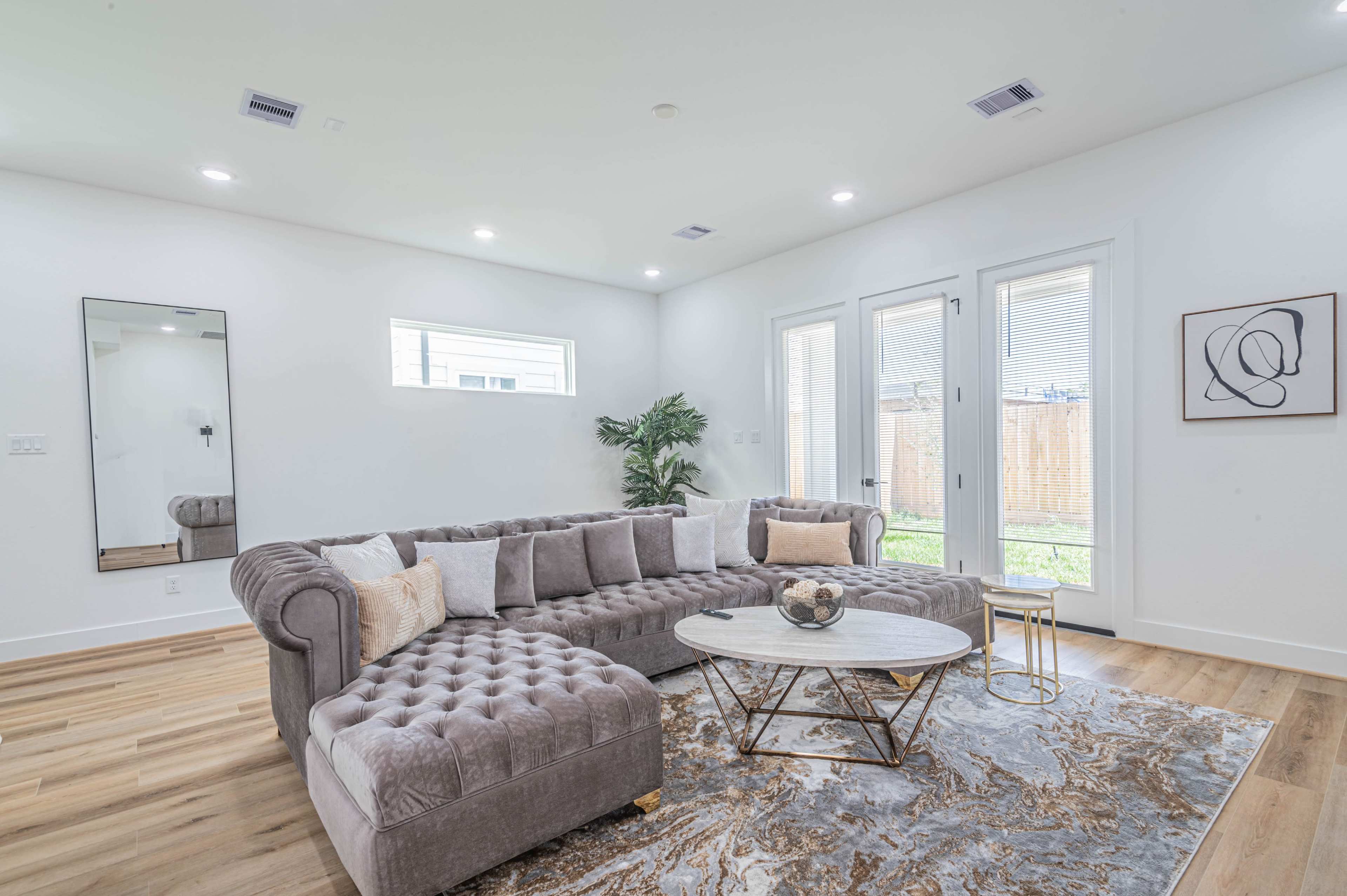 The living room features a gray tufted sectional sofa, a round coffee table, and large windows that open to a backyard.