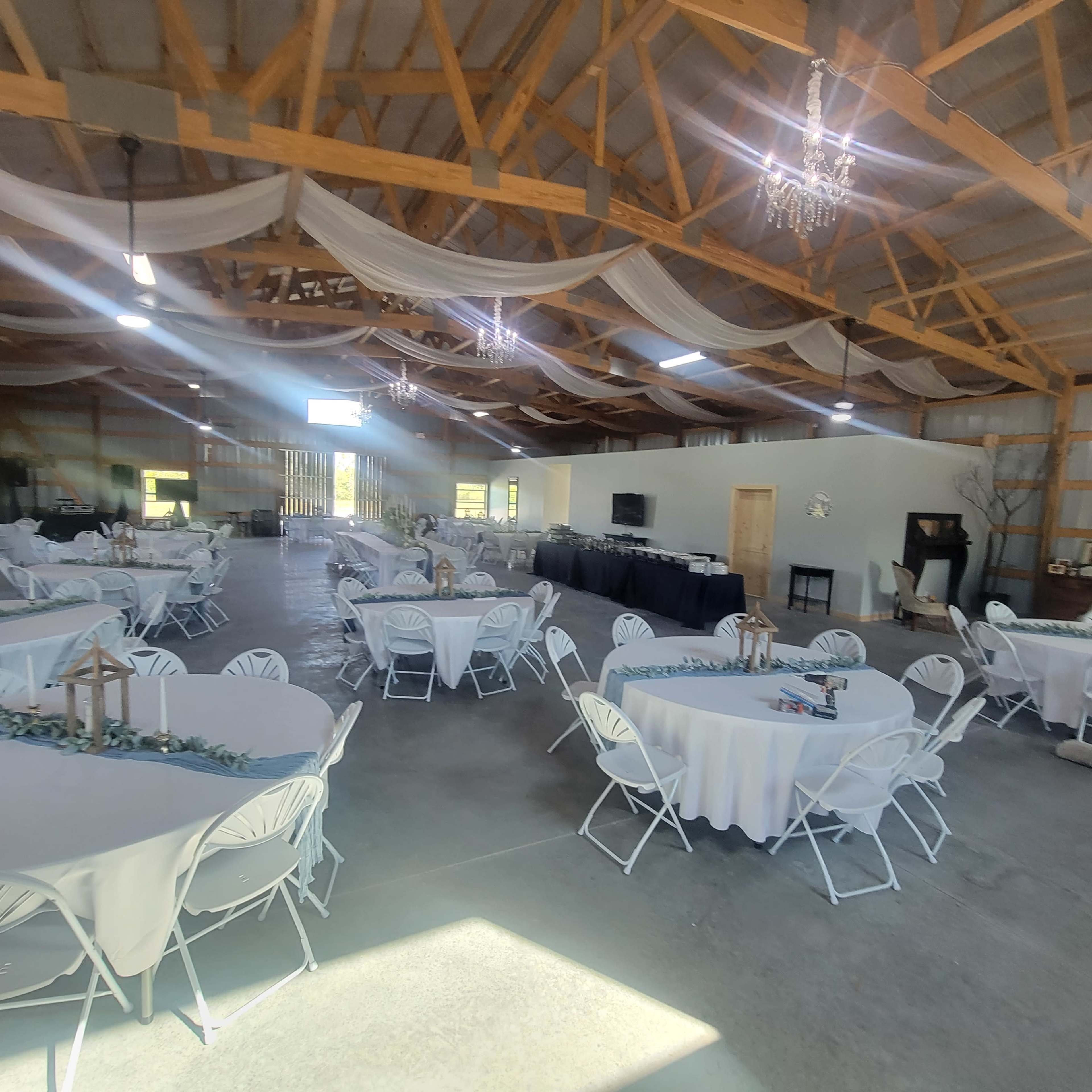 A large, open event space with round tables covered in white tablecloths, arranged neatly under a wooden ceiling with chandeliers.