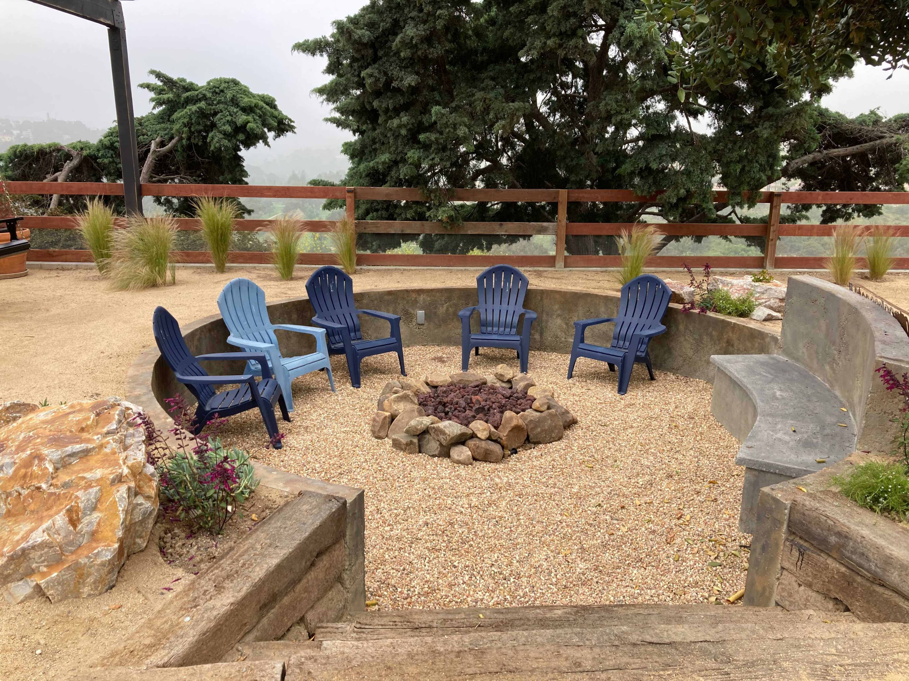 A circular seating area surrounded by gravel features five blue Adirondack chairs arranged around a stone fire pit.