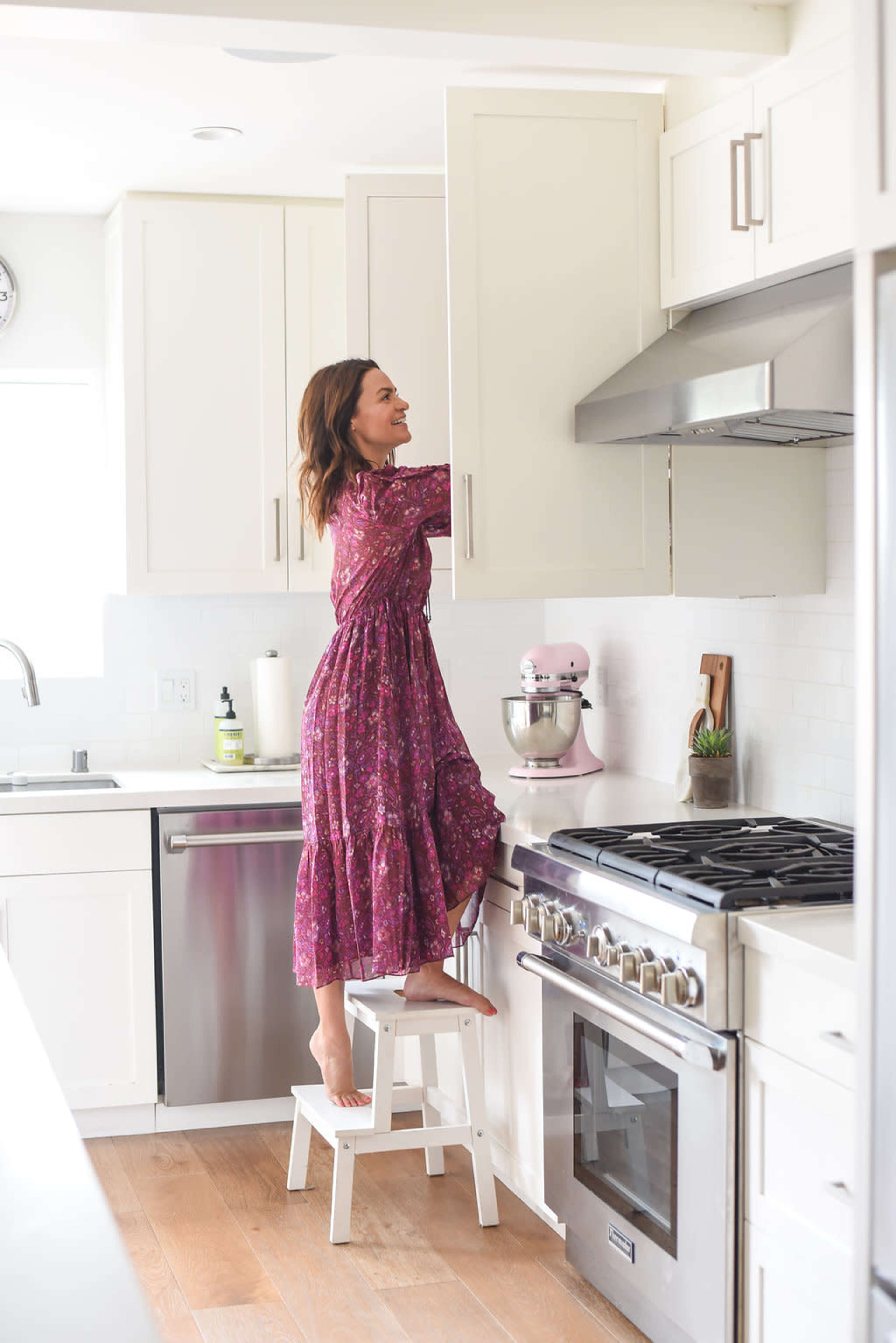 A woman in a purple dress stands on a stool to reach for an item in a kitchen cabinet.