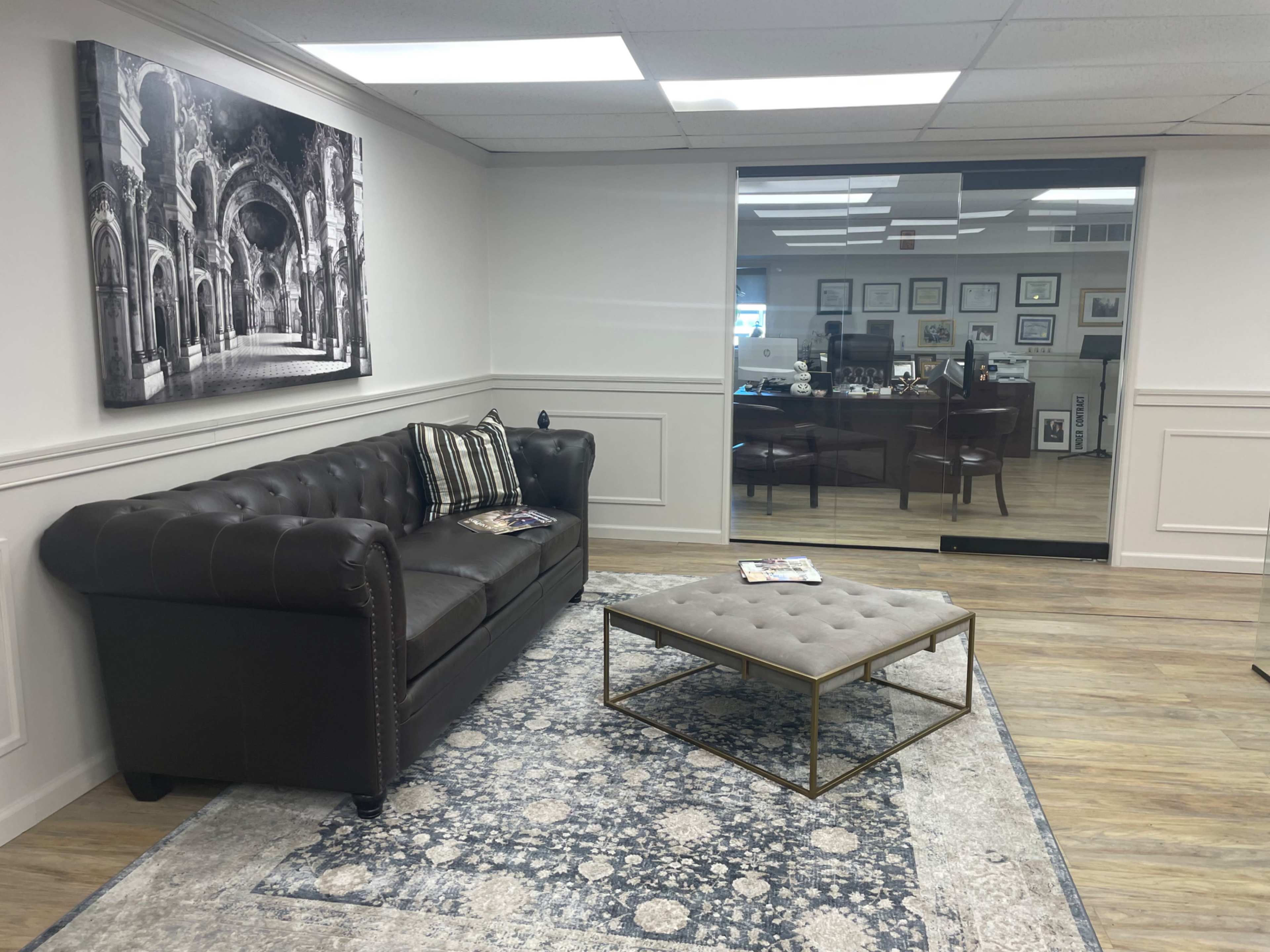 A waiting area featuring a brown tufted leather sofa, a coffee table on a decorative rug, and a wall with a black-and-white photograph.