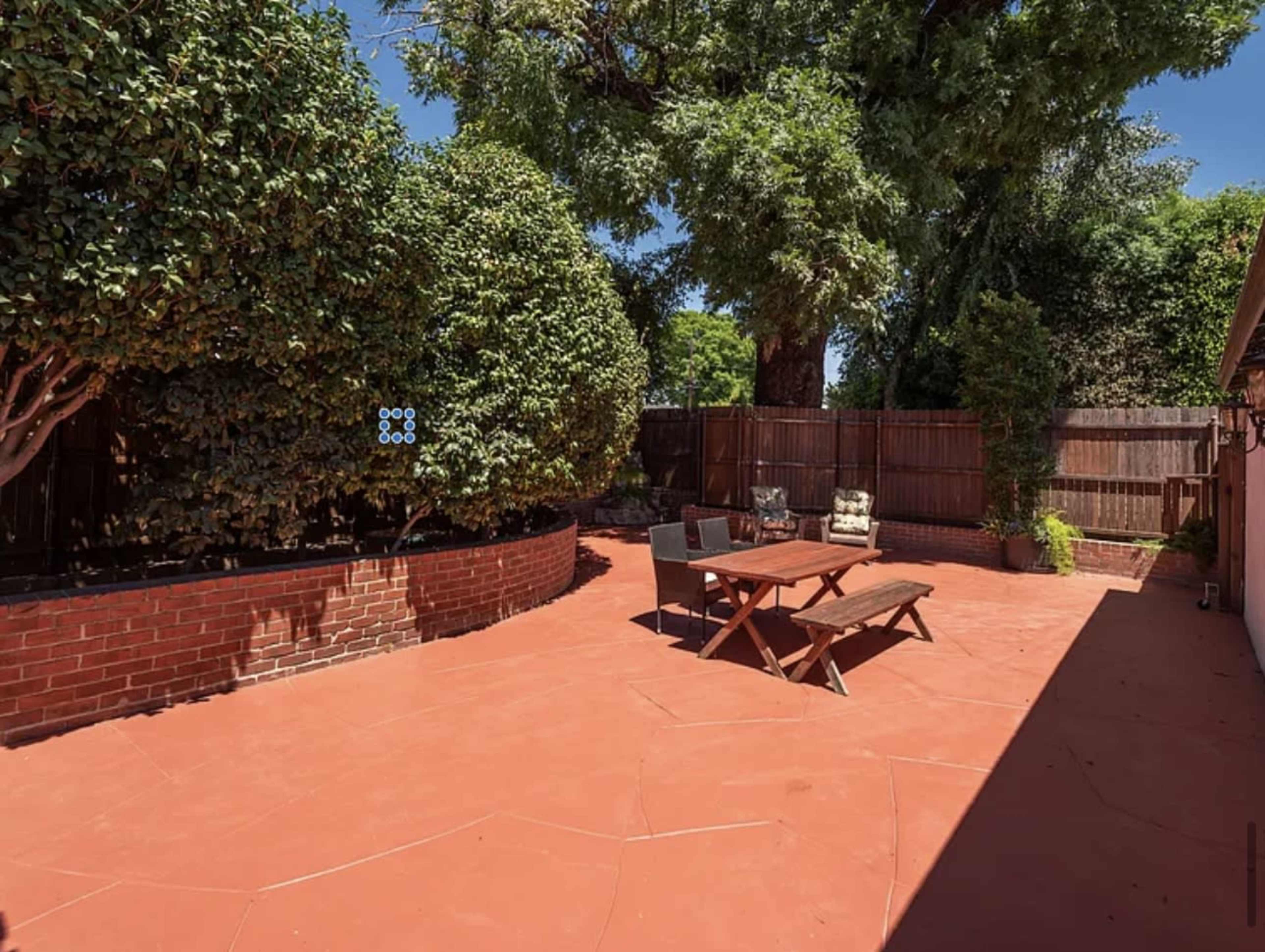 The image shows a red-tiled backyard area with several trees, a brick border, and a wooden picnic table surrounded by chairs.