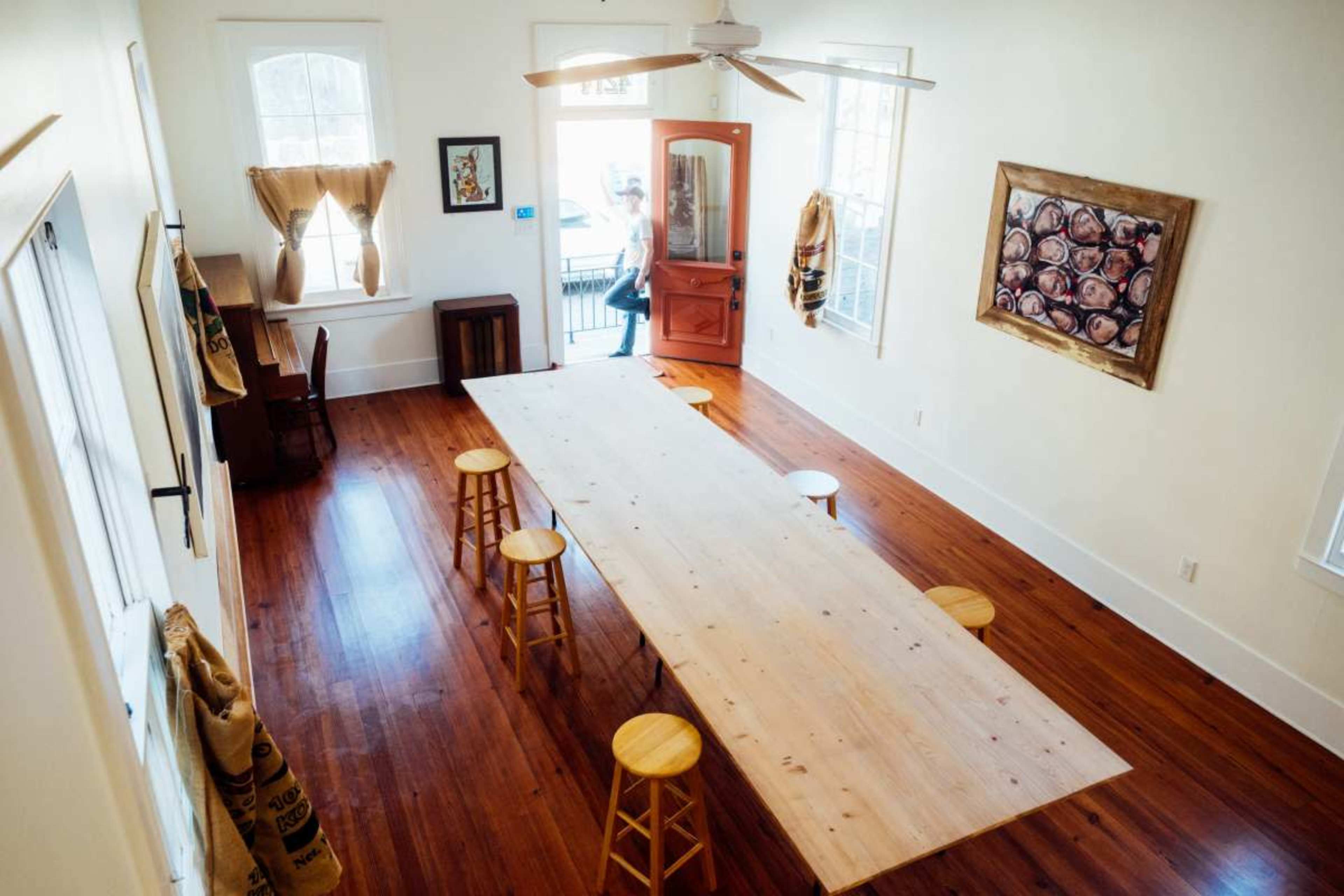 A spacious room features a long wooden table surrounded by several stools, with large windows and a door leading outside.