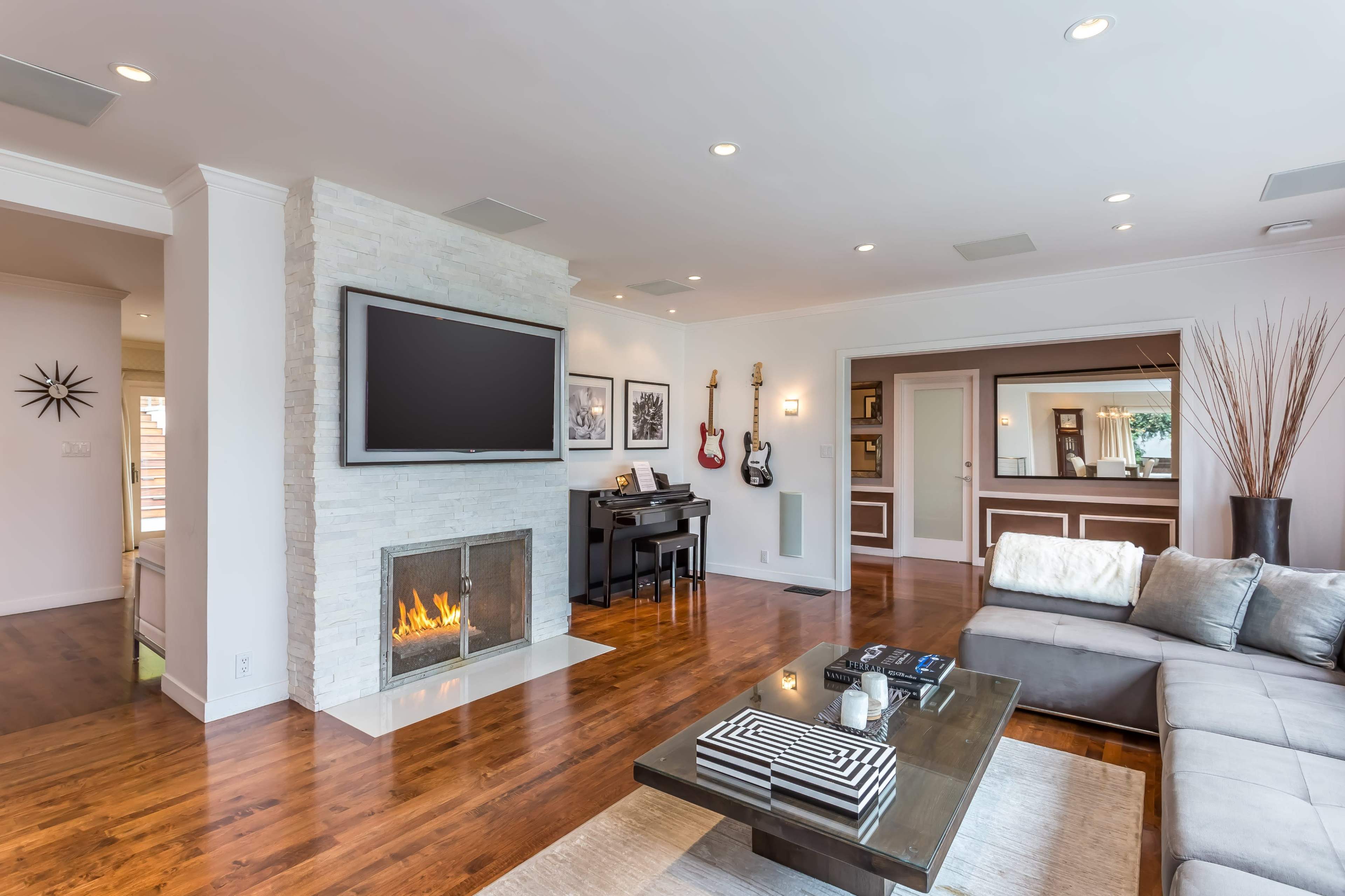 The image shows a modern living room with a flat-screen TV mounted above a stone fireplace, a piano, and a sectional couch on hardwood floors.