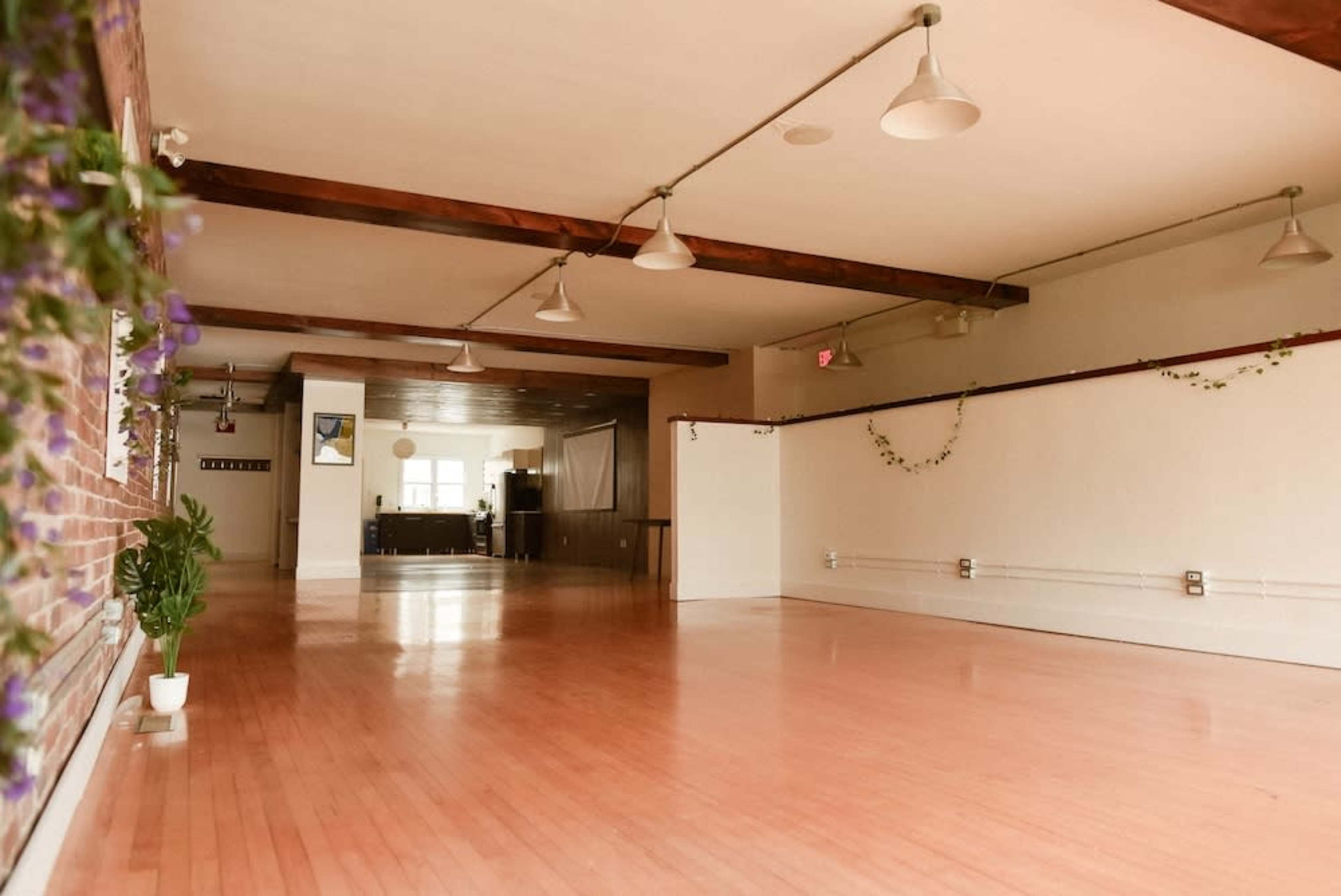 The image shows a spacious, well-lit room with wooden floors, exposed beams, and minimal decorations along the walls.