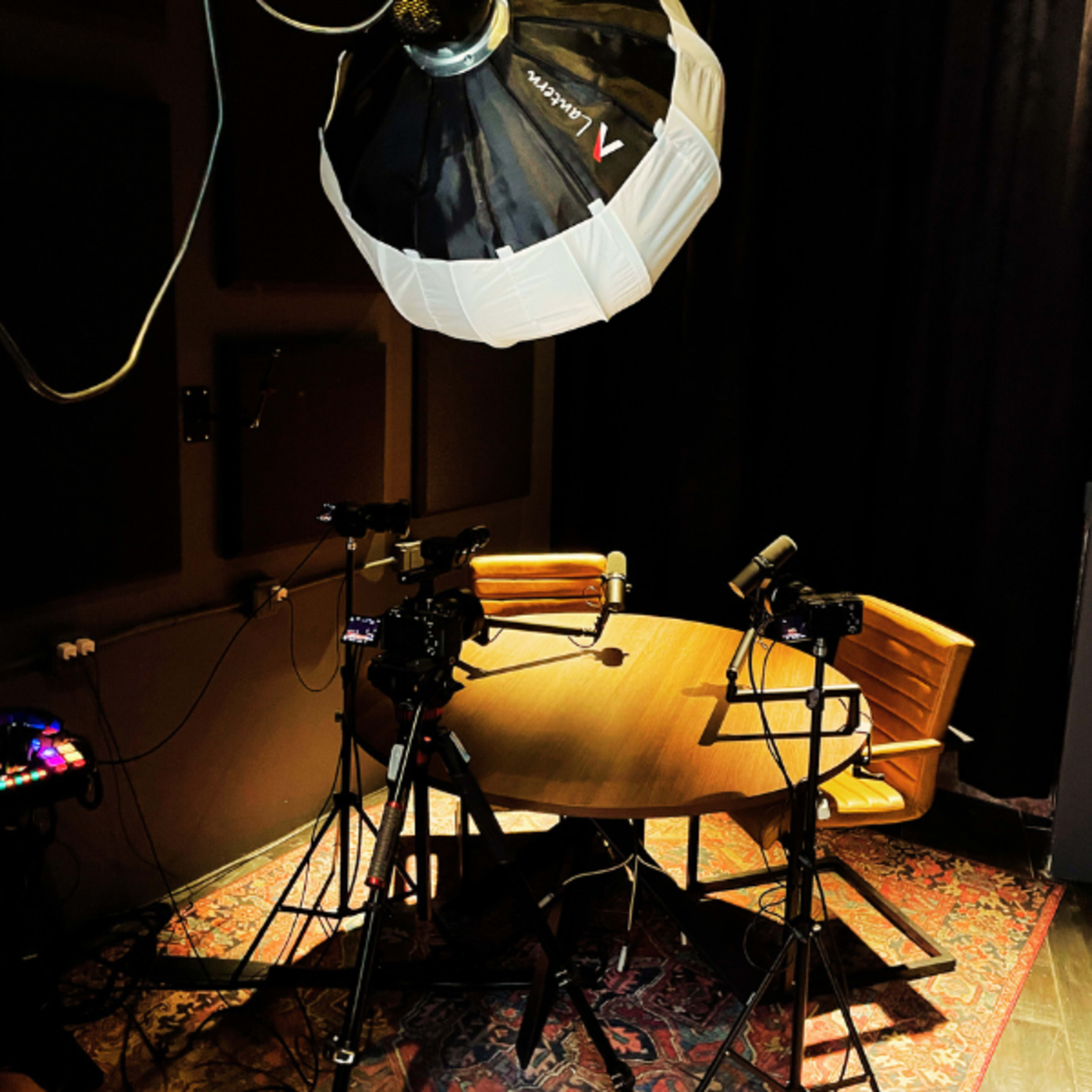 The image shows a small studio setup featuring a round table with two chairs, surrounded by camera tripods and lighting equipment.