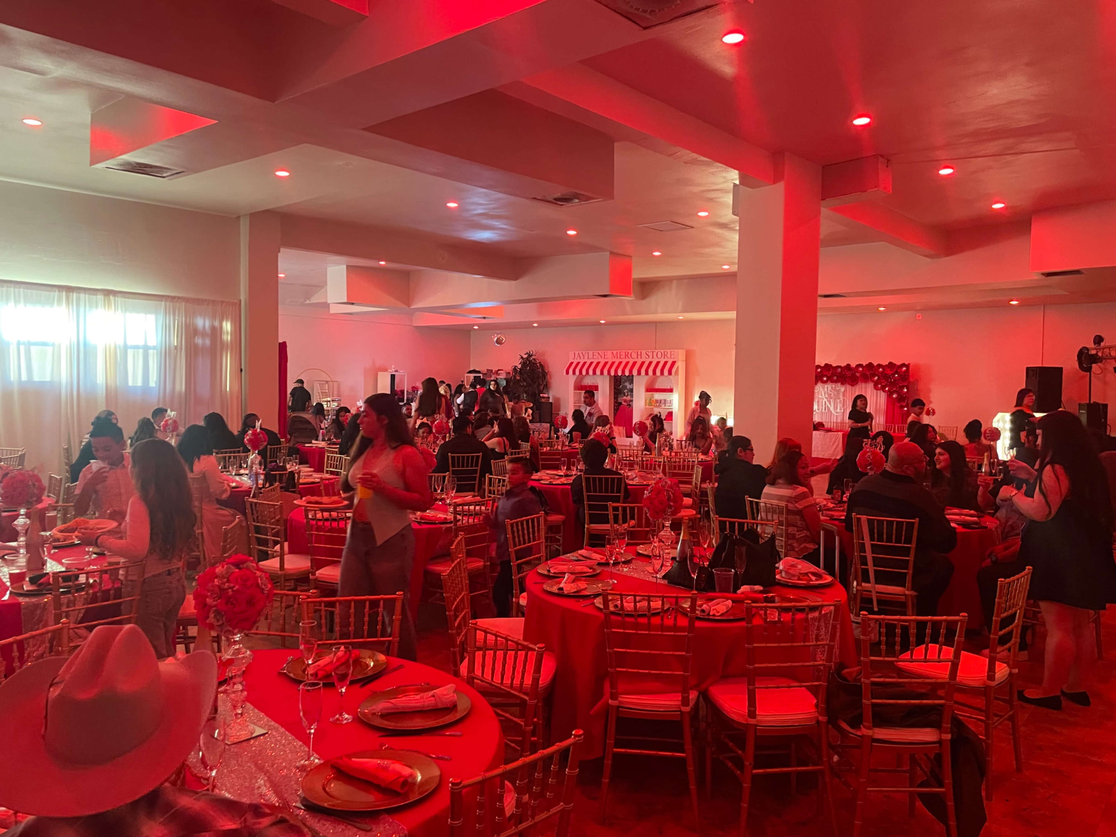 A banquet hall filled with guests seated at elegantly set tables, illuminated by red lighting and decorated with floral centerpieces.