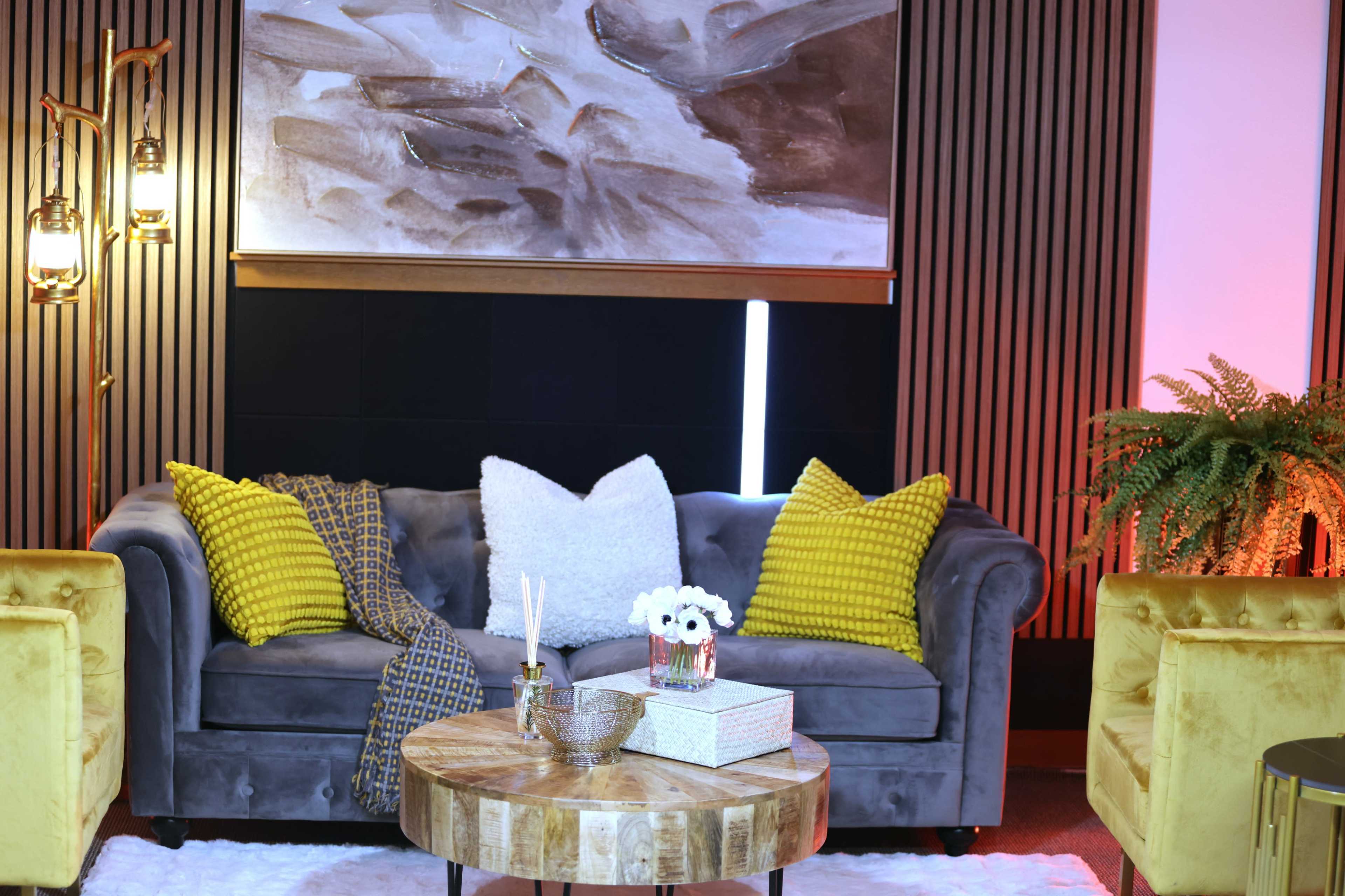 The image shows a cozy living room setup featuring a gray couch adorned with yellow and white pillows, a round wooden coffee table, and decorative plants, all set against a backdrop of textured walls and ambient lighting.