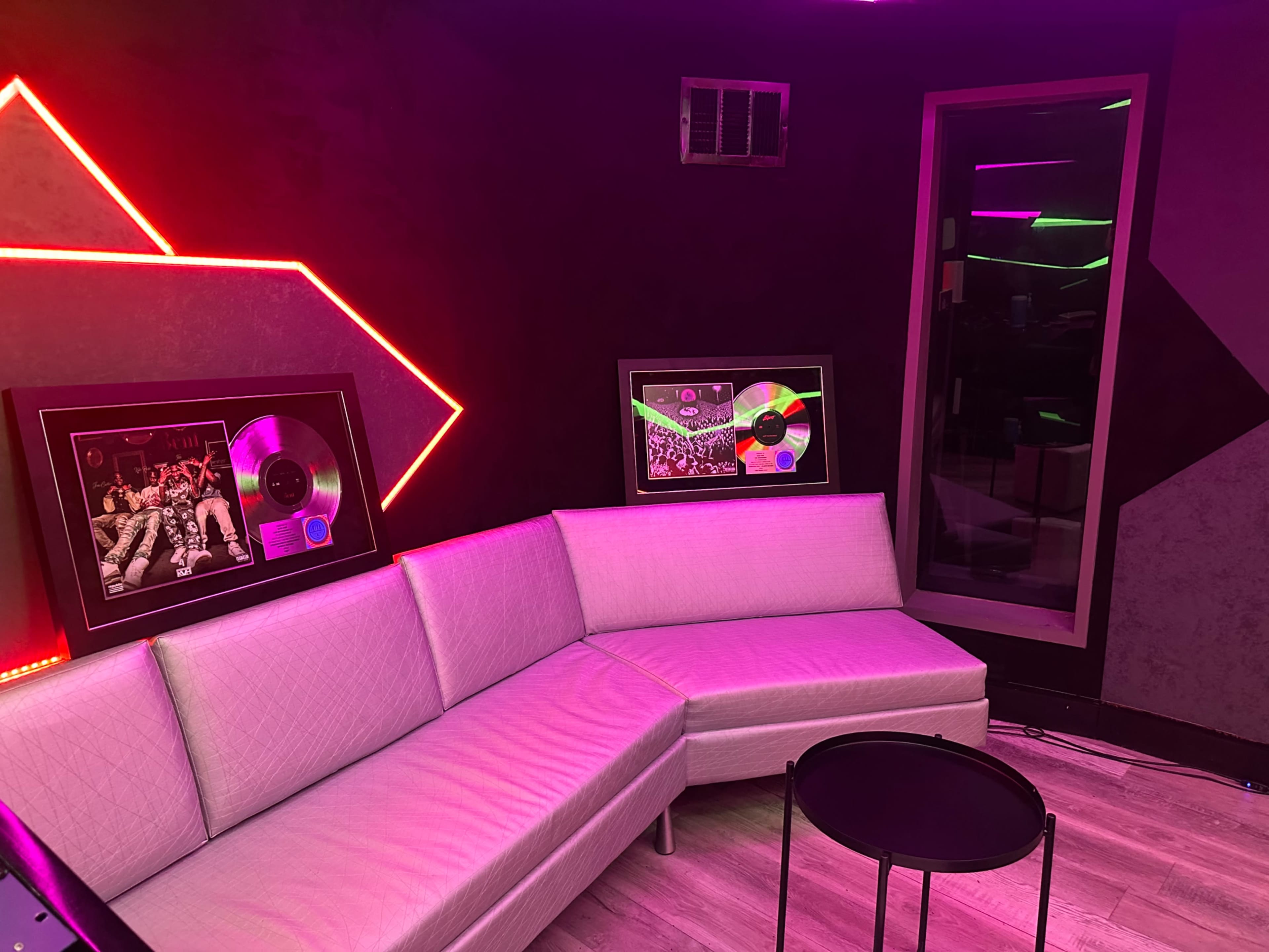 The image shows a modern lounge area with a white corner sofa, a small black table, and framed music awards on the walls illuminated by colorful neon lighting.