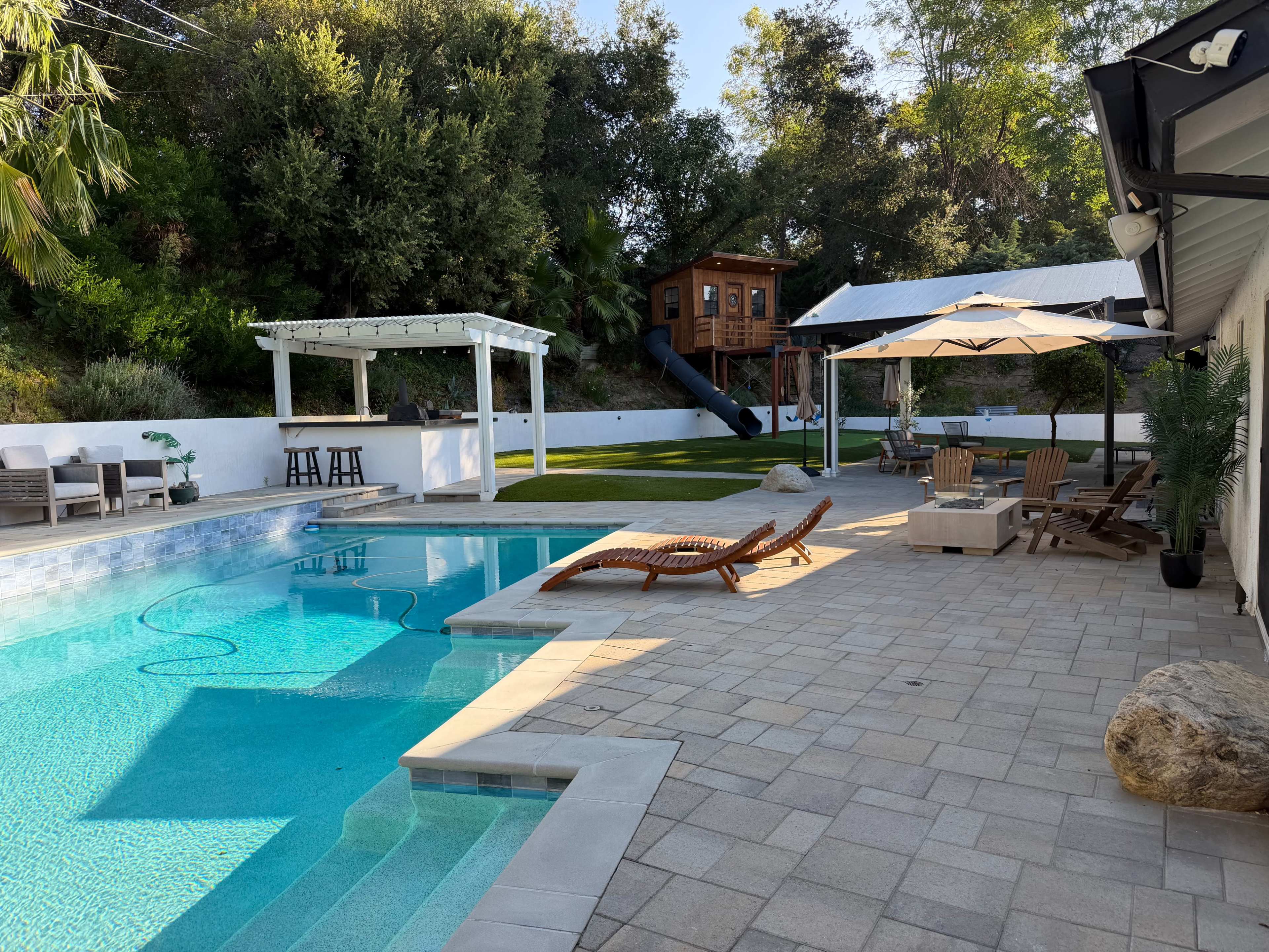 The image shows a tranquil outdoor space featuring a swimming pool, a patio with lounge chairs, a covered sitting area, and a wooden playhouse with a slide in the background.