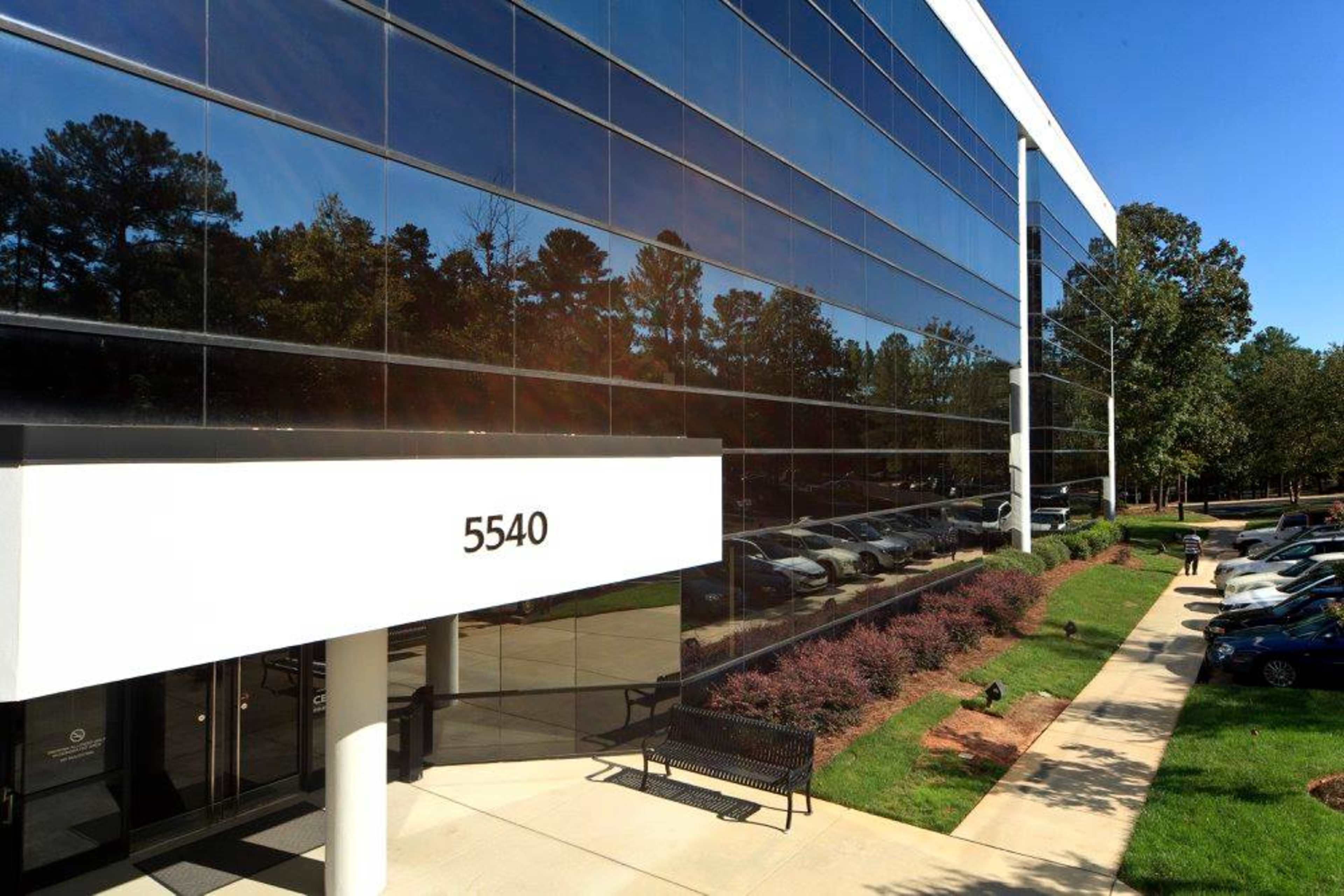 A modern building with large glass windows reflecting surrounding trees and a pathway leading to the entrance, labeled "5540," along with parked cars nearby.