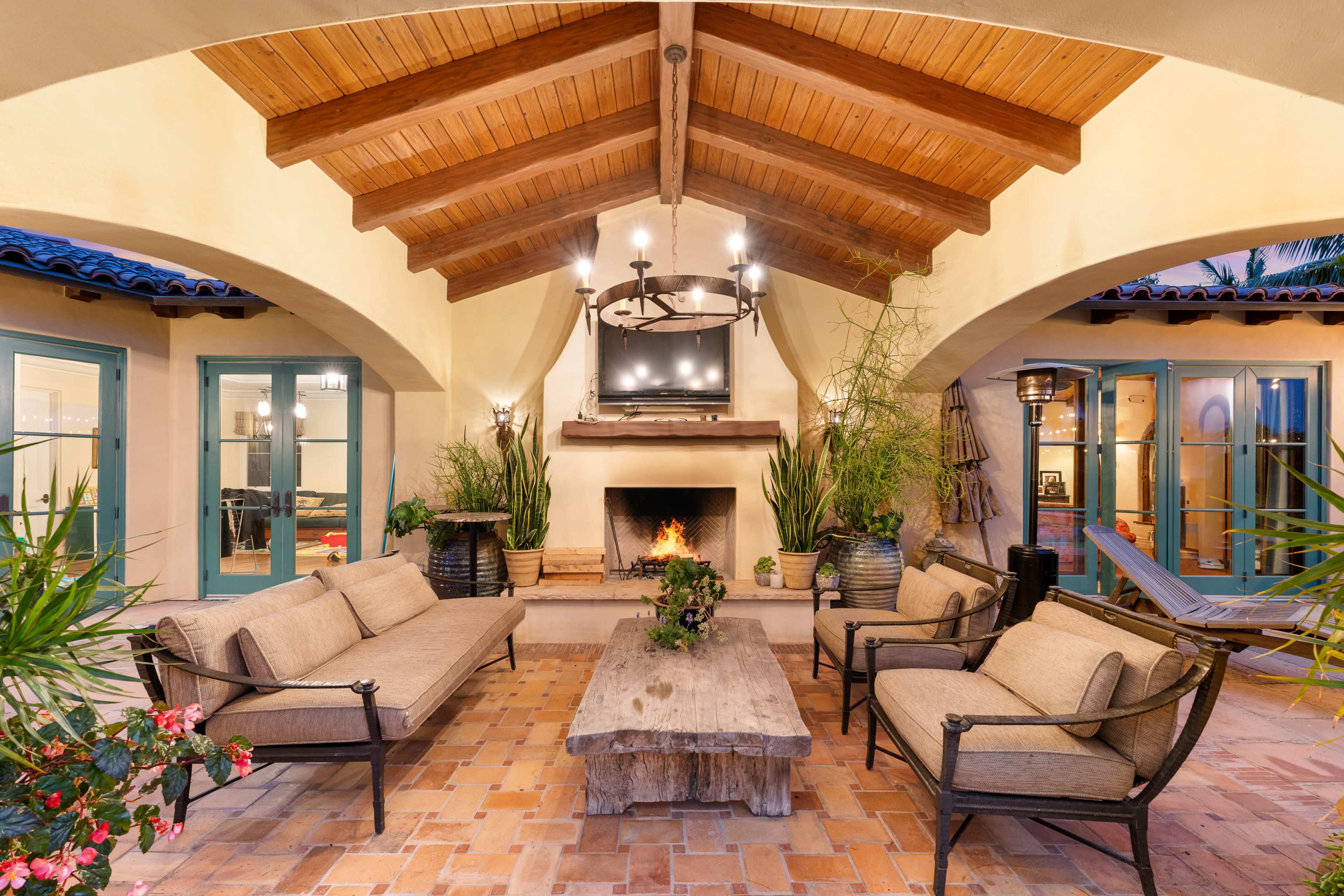 A cozy outdoor living space with a fireplace, wooden beams, and comfortable seating surrounded by potted plants.