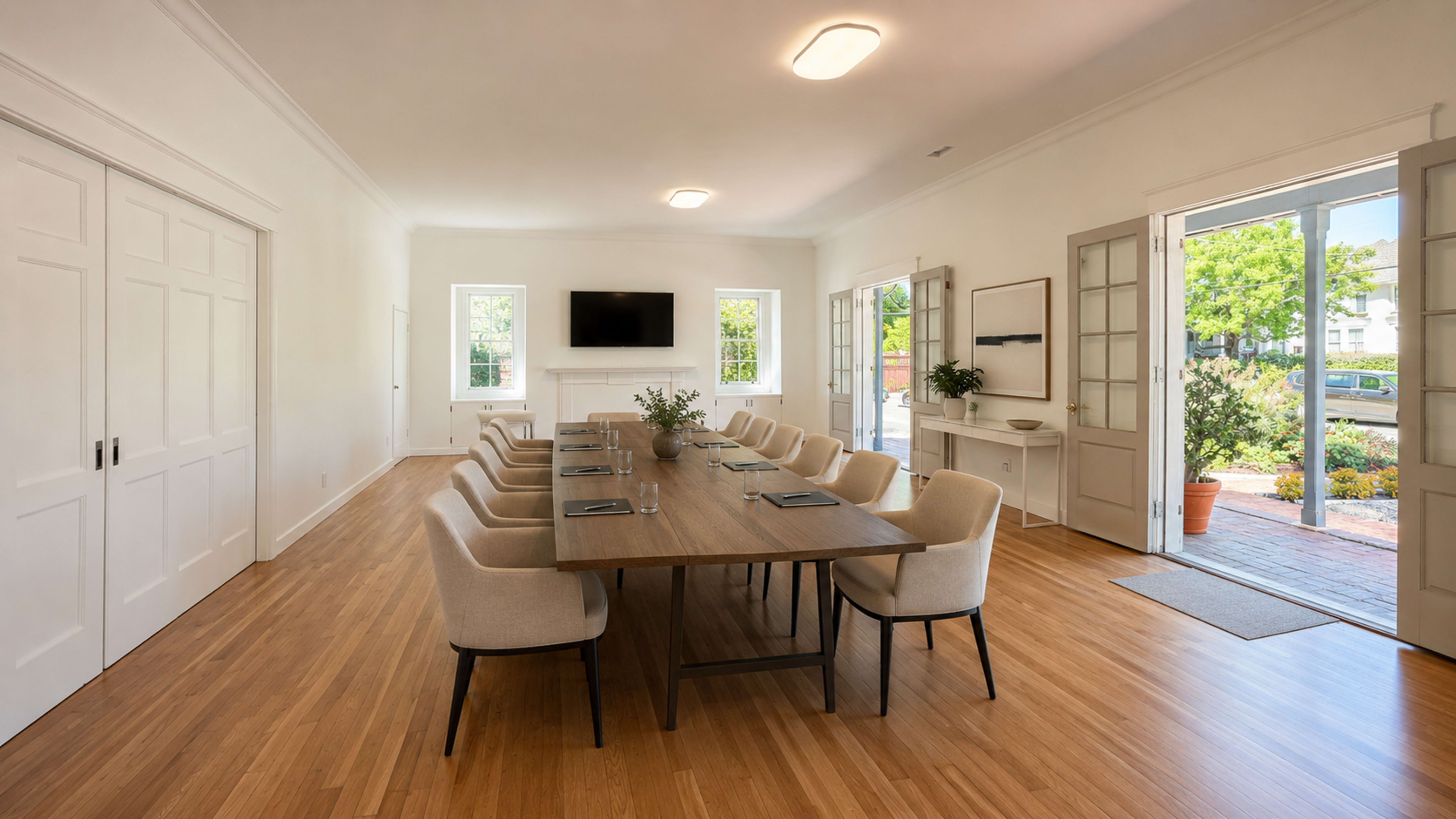 A spacious meeting room features a long wooden table surrounded by beige chairs, with a television mounted on the wall and sunlight streaming through open doors.