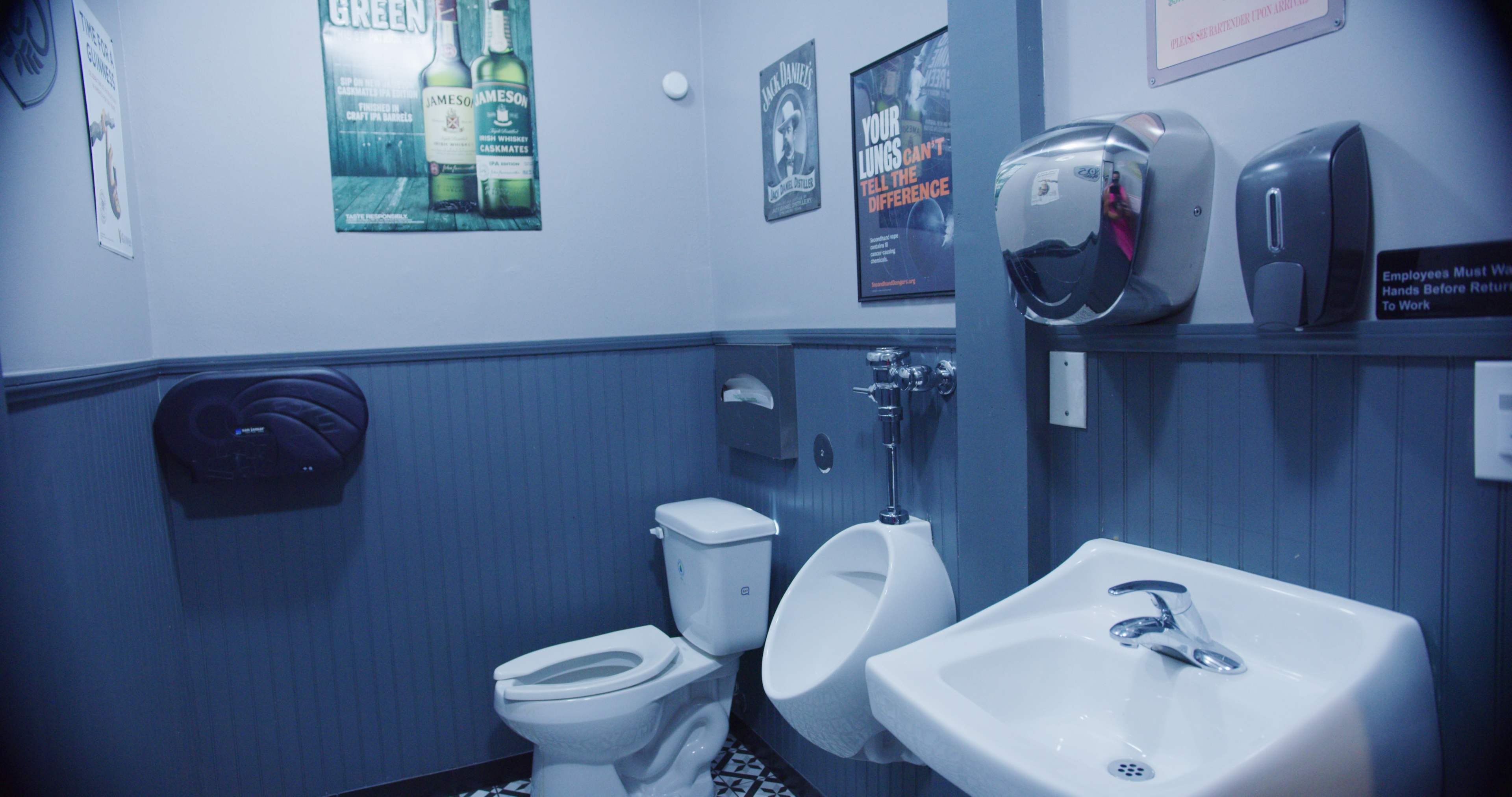 A small, modern restroom with a toilet, urinal, sink, and various wall posters.