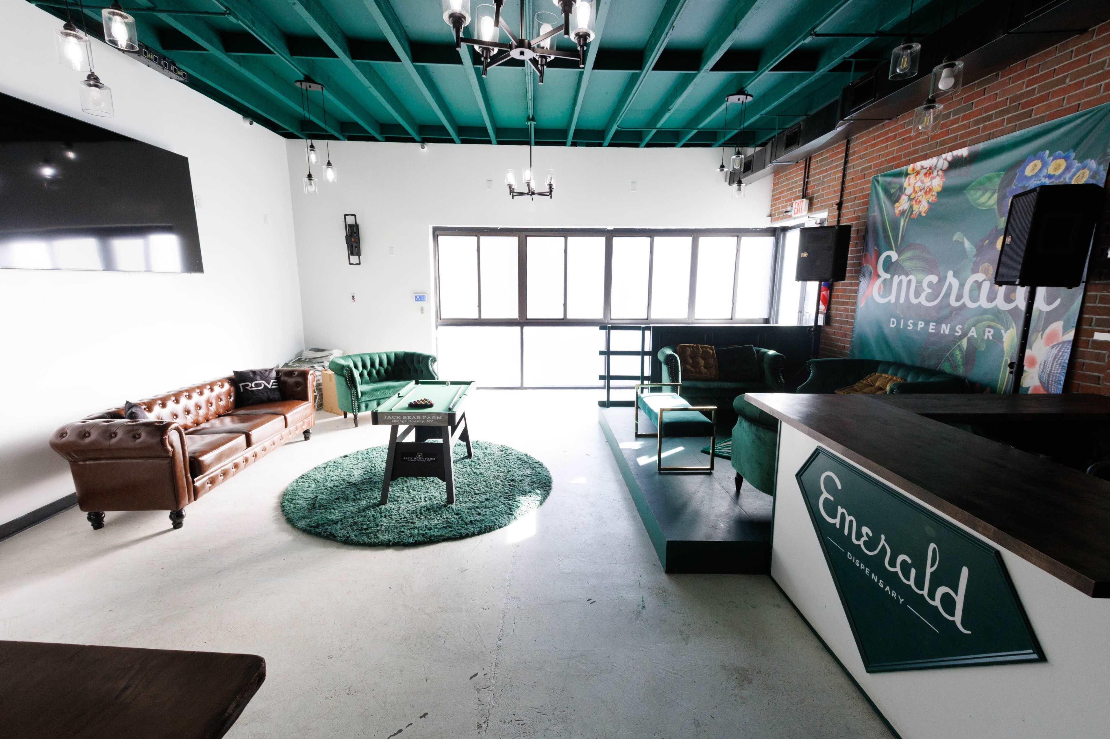 The interior of a modern dispensary features a green ceiling, two sofas, a coffee table on a circular rug, and a reception area with branding visible on the counter.