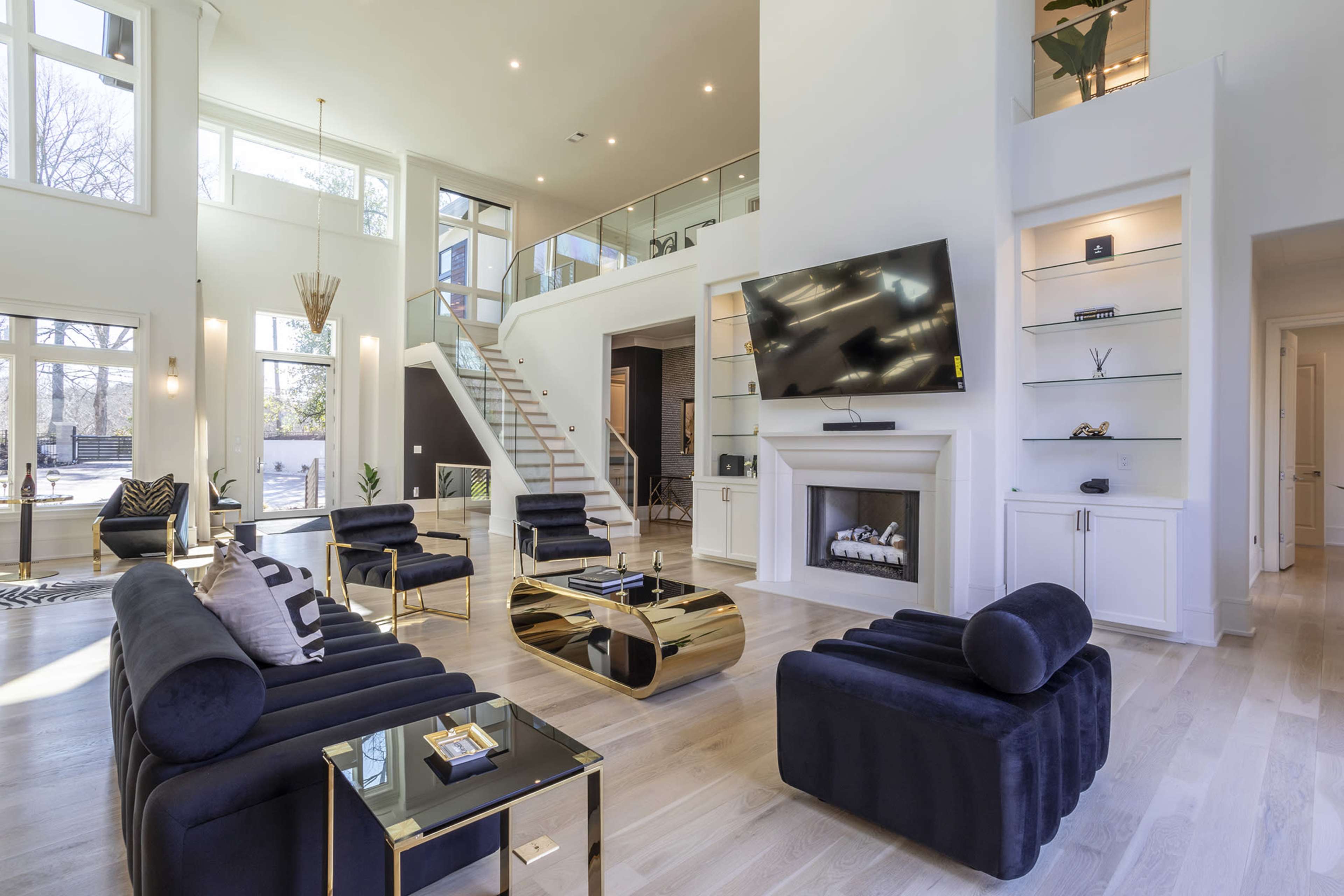 The spacious living room features large windows, a modern television mounted above a fireplace, and sleek black seating arranged around a gold coffee table.