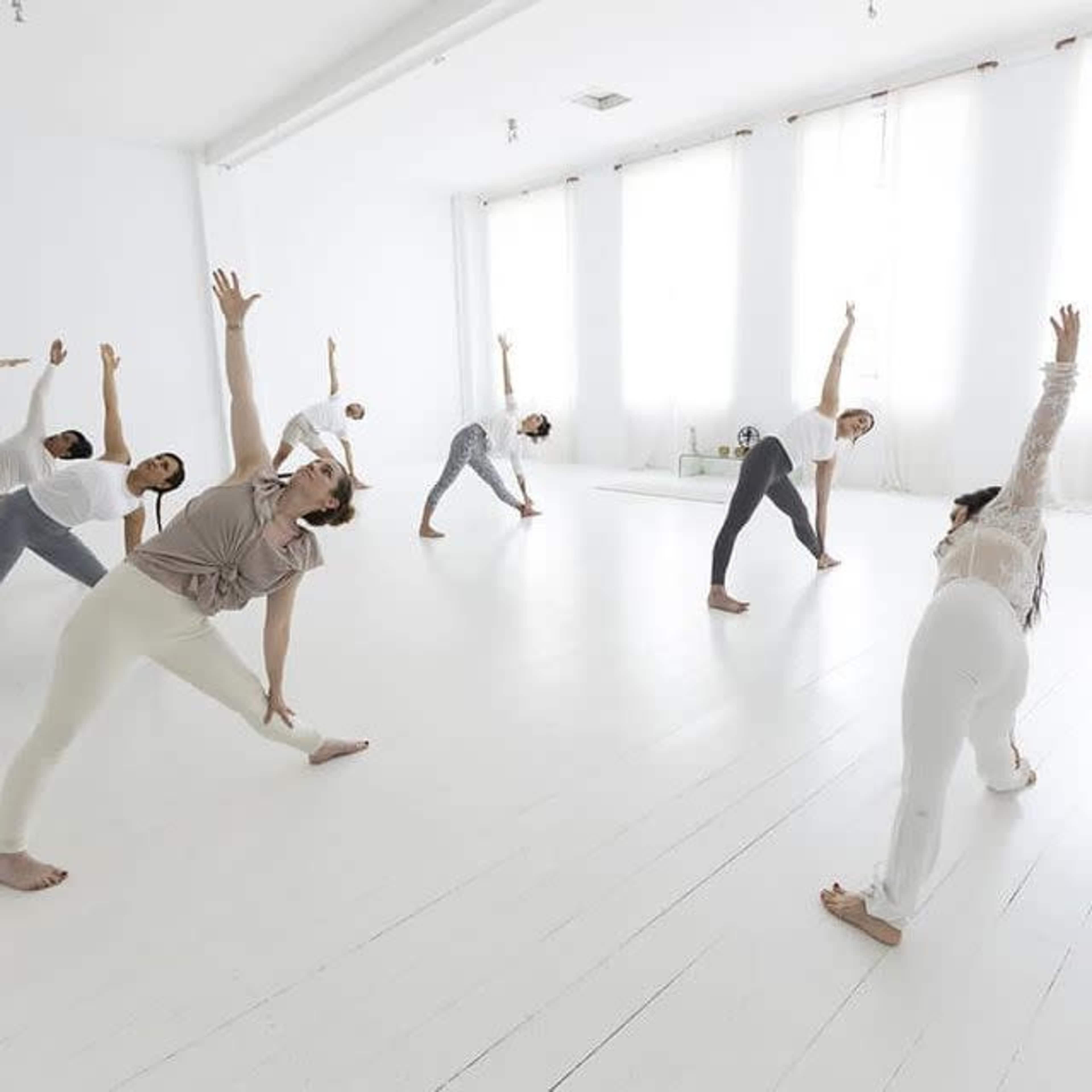 A group of individuals is engaged in a yoga or stretching session inside a spacious, well-lit studio with white walls and large windows.