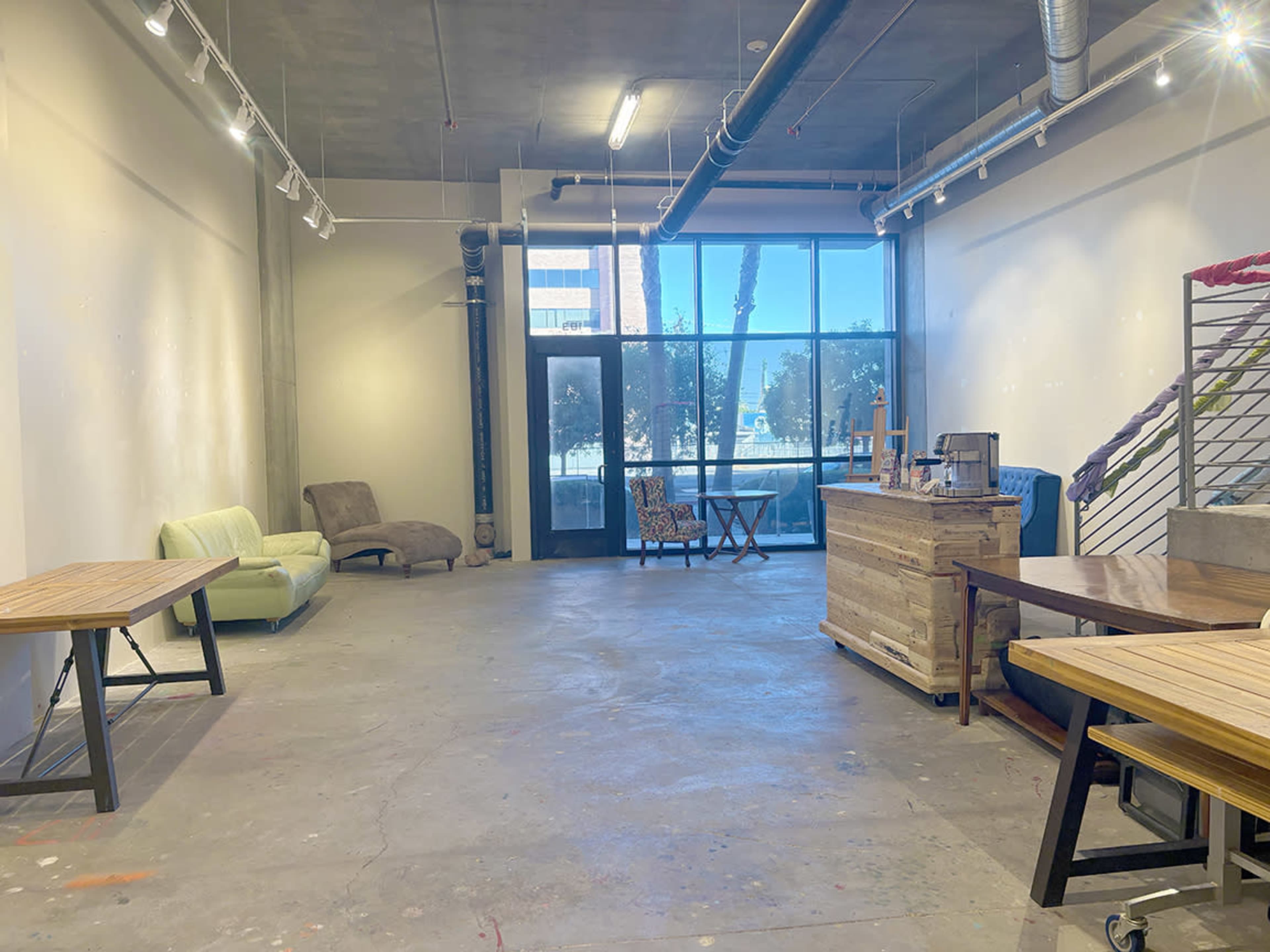 The image shows an empty interior space with concrete flooring, large windows, and various wooden furniture pieces including tables and a couch.