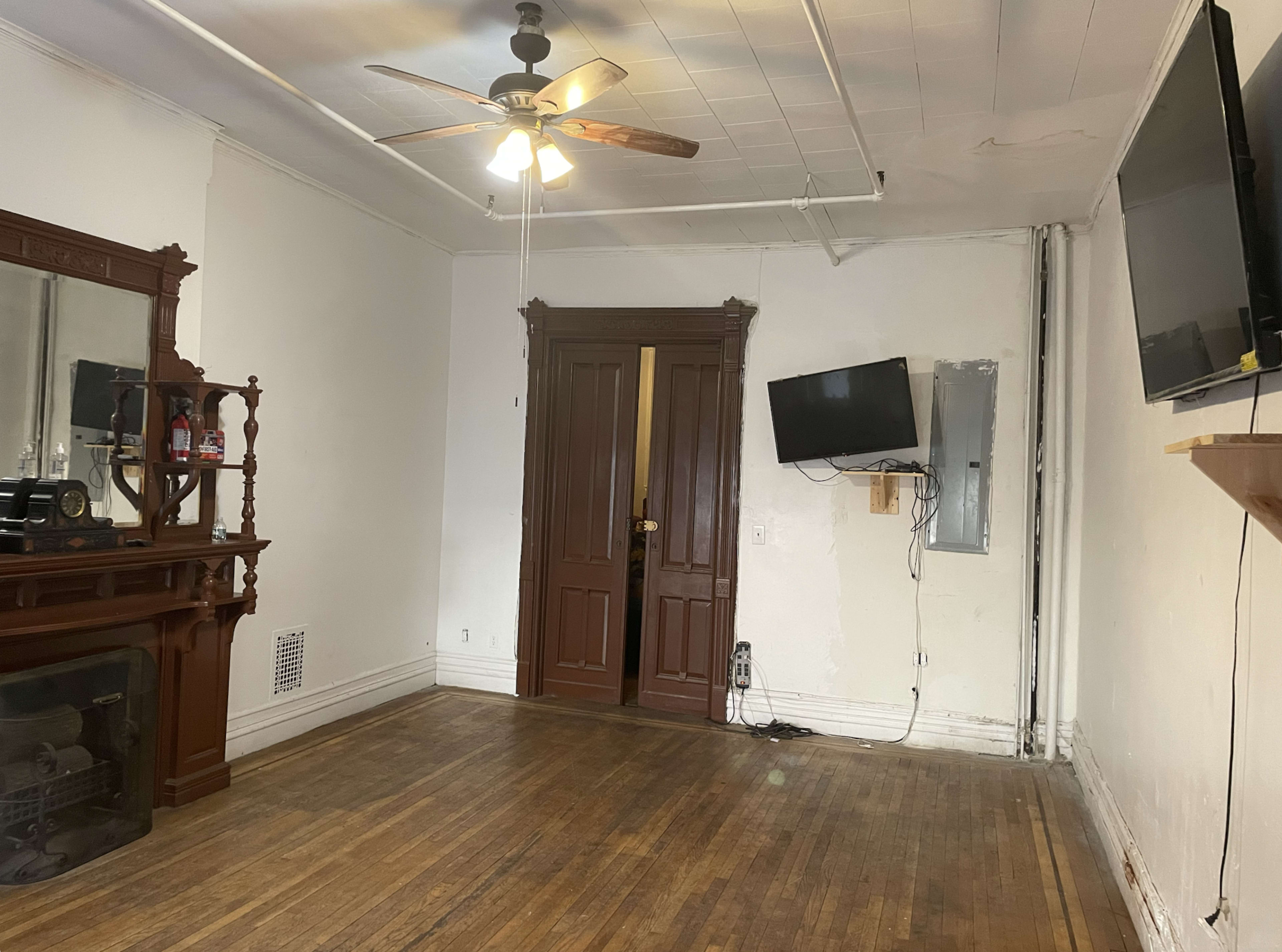 The image shows a room with wooden flooring, a ceiling fan, and a wall-mounted television, featuring a closed door with a decorative frame.