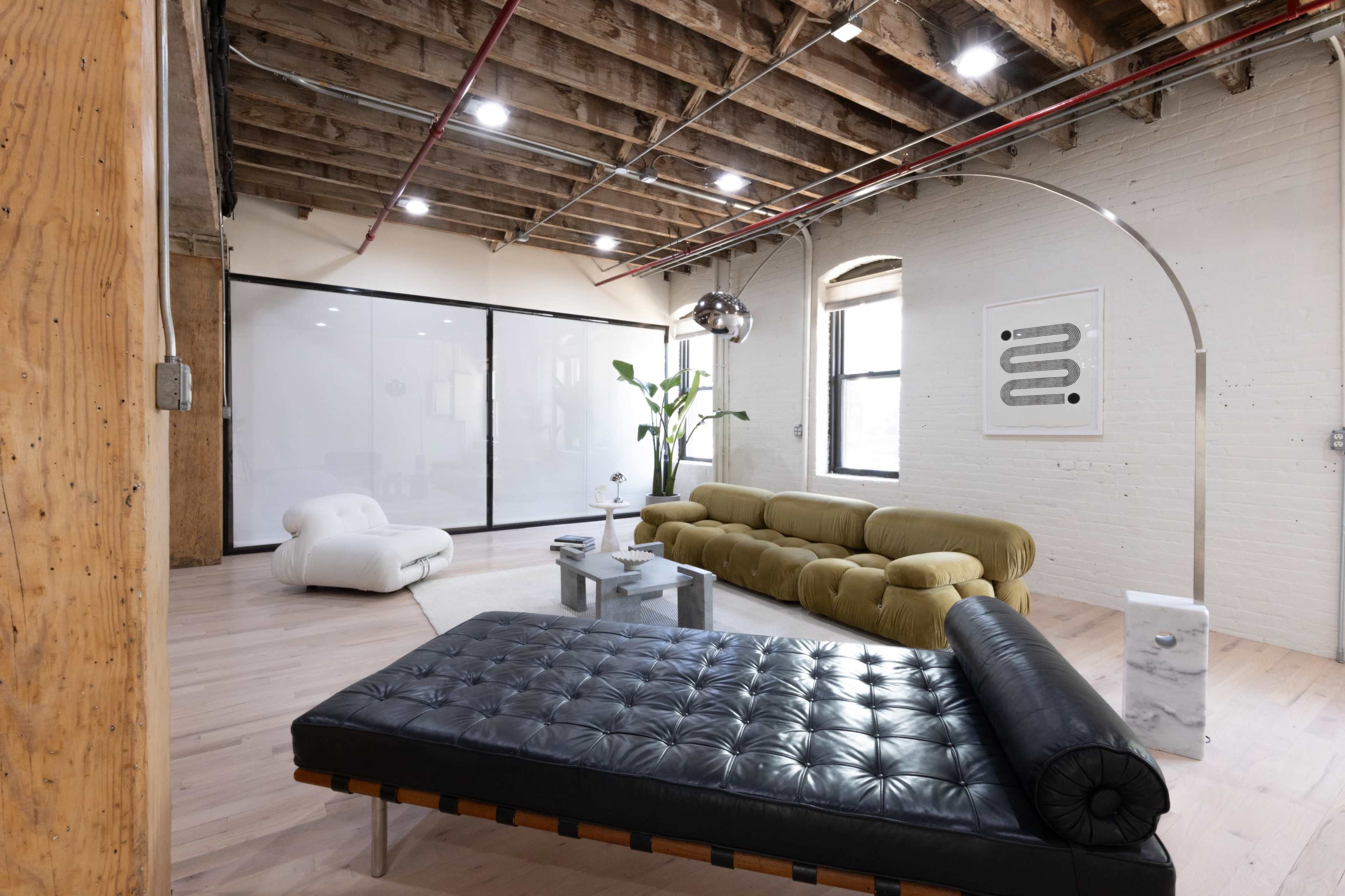 The image shows a spacious, modern living room with exposed wooden beams, a green sofa, a black chaise lounge, and large windows allowing natural light.