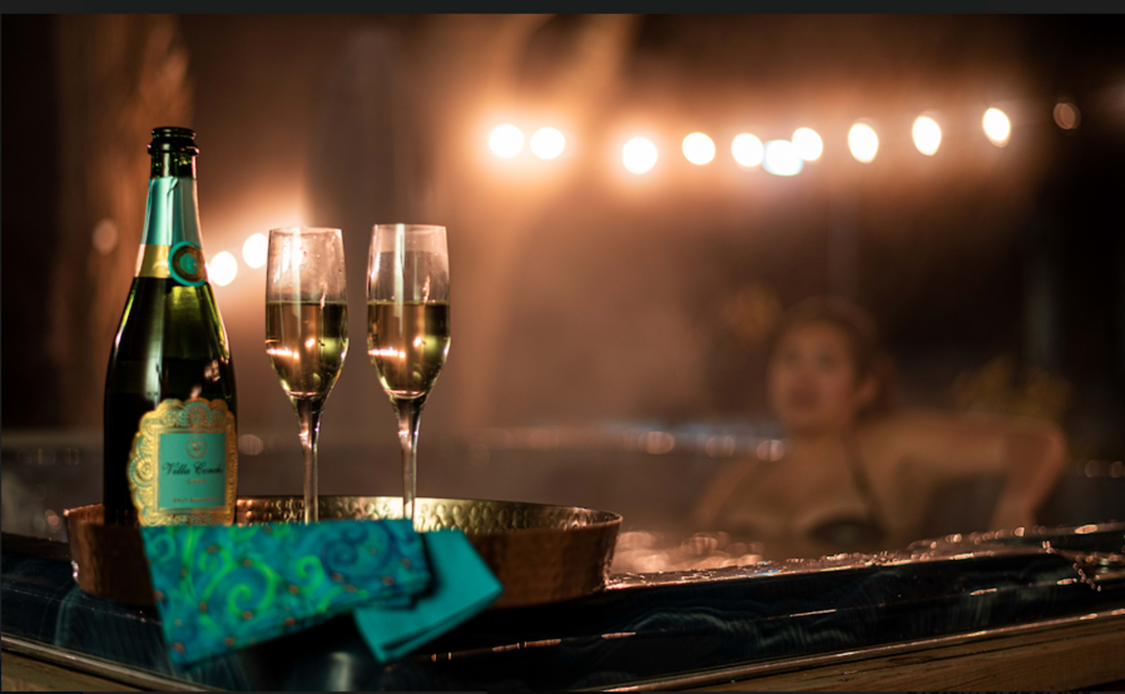 A bottle of sparkling wine and two fluted glasses are set on a tray beside a hot tub, with soft lighting in the background.