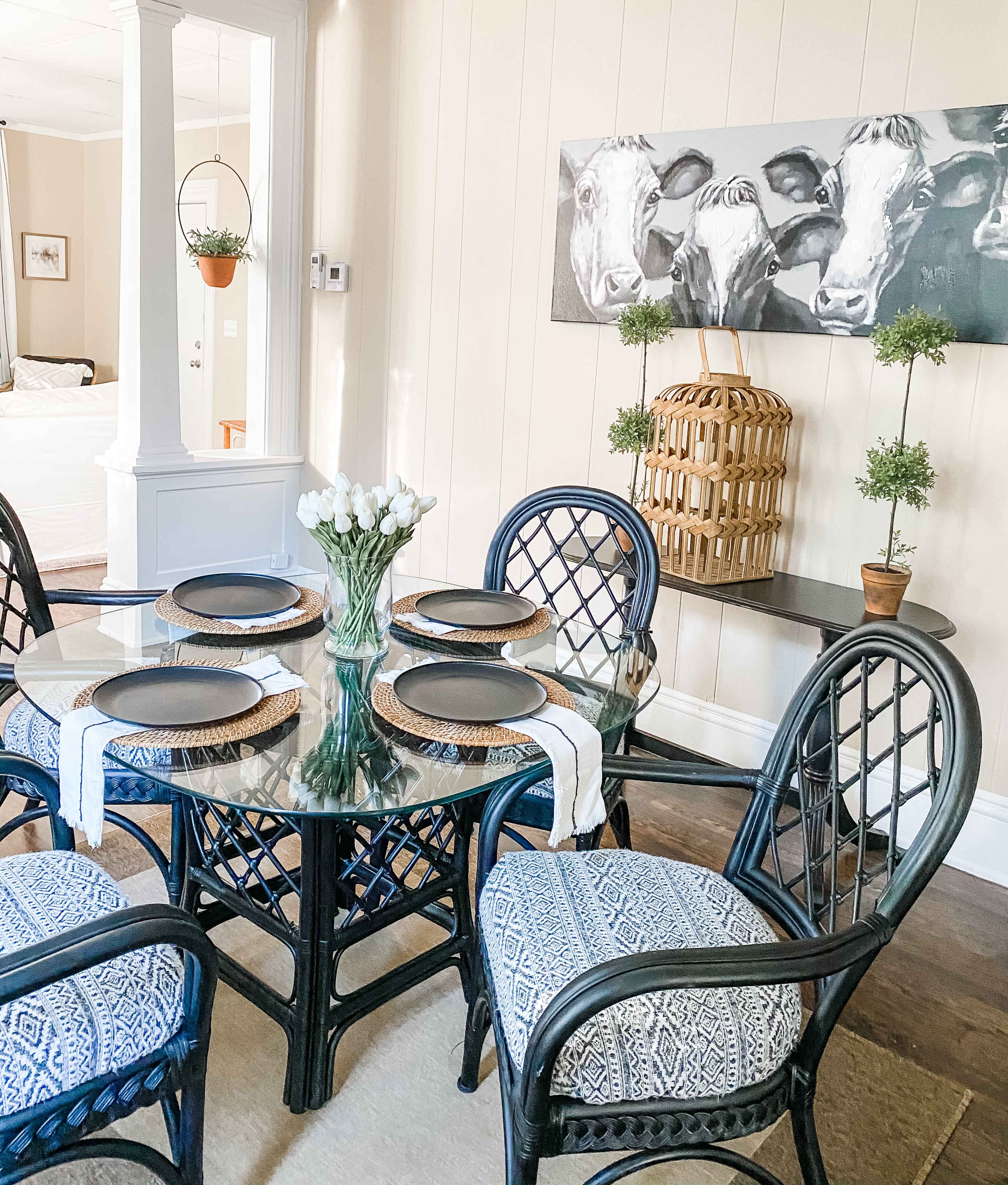 A dining room features a round glass table surrounded by six patterned chairs, with a vase of tulips at the center and a wall display of cow portraits above a wooden shelf.