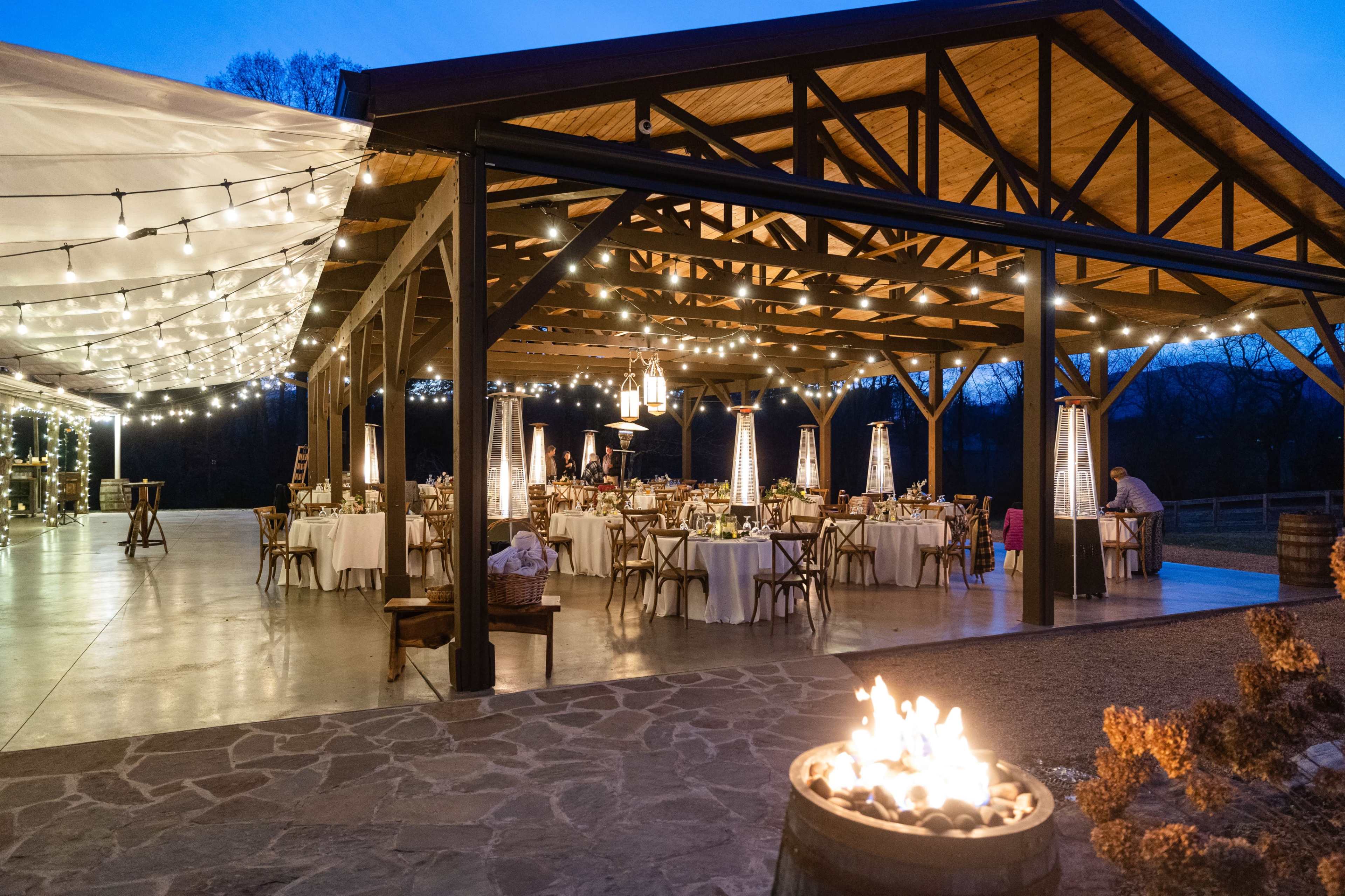 A spacious outdoor event venue features neatly arranged round tables under string lights and a nearby fire pit.