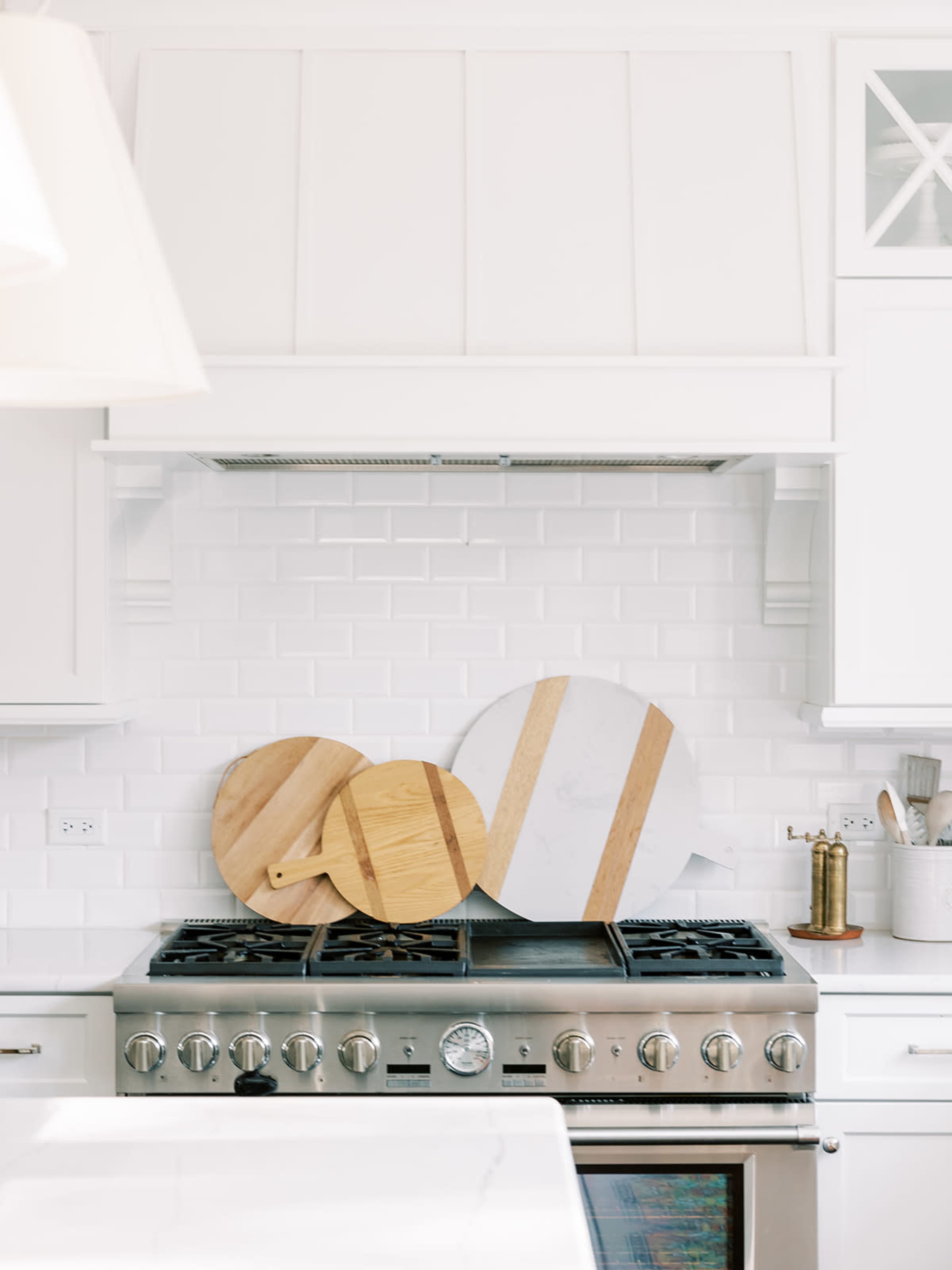 Three cutting boards of varying sizes, two made of wood and one of marble, are propped against the backsplash of a kitchen stove in a white kitchen.