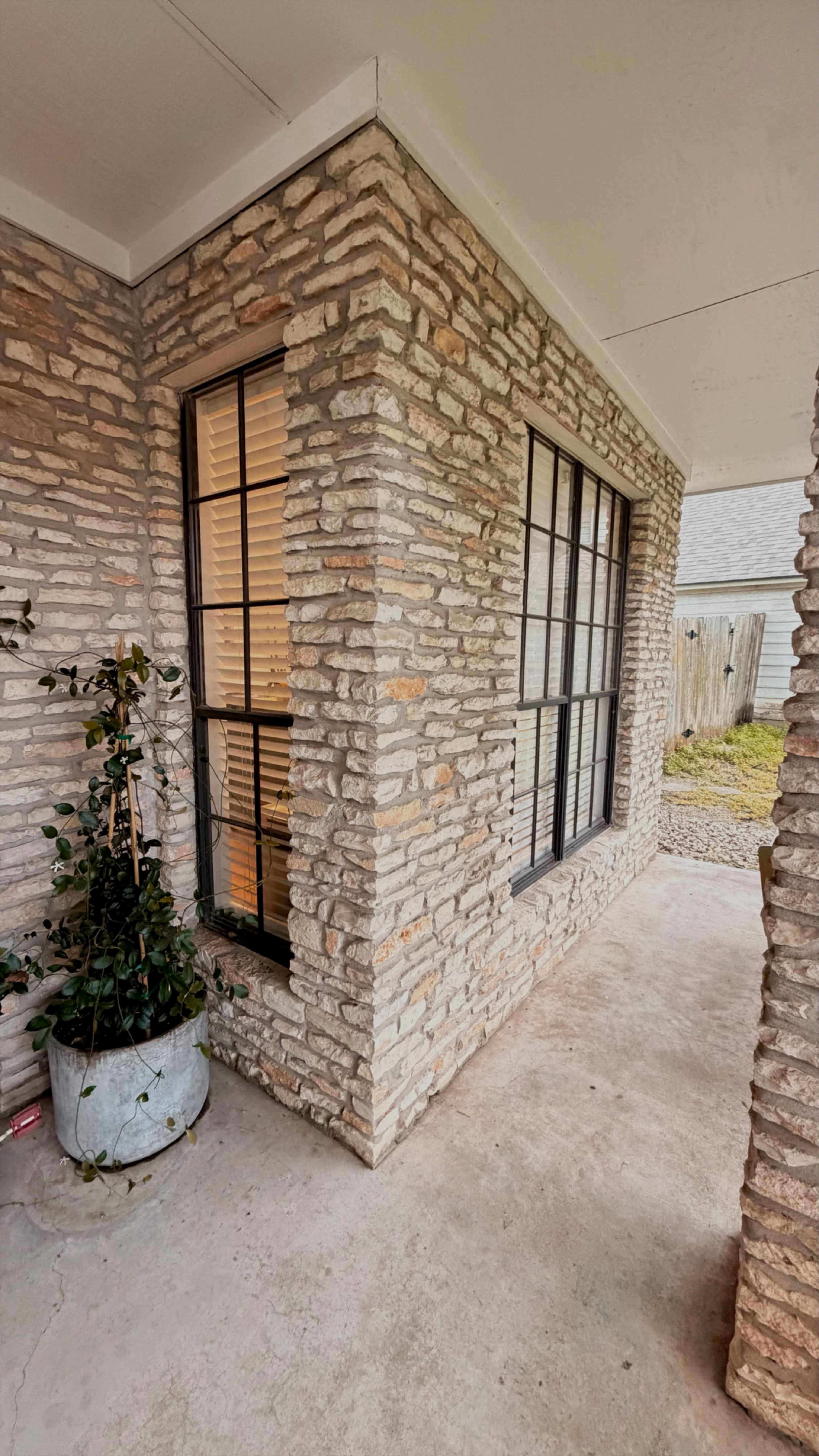 The image shows a corner of a house featuring a stone facade, two windows with black frames, and a potted plant beside the entrance.