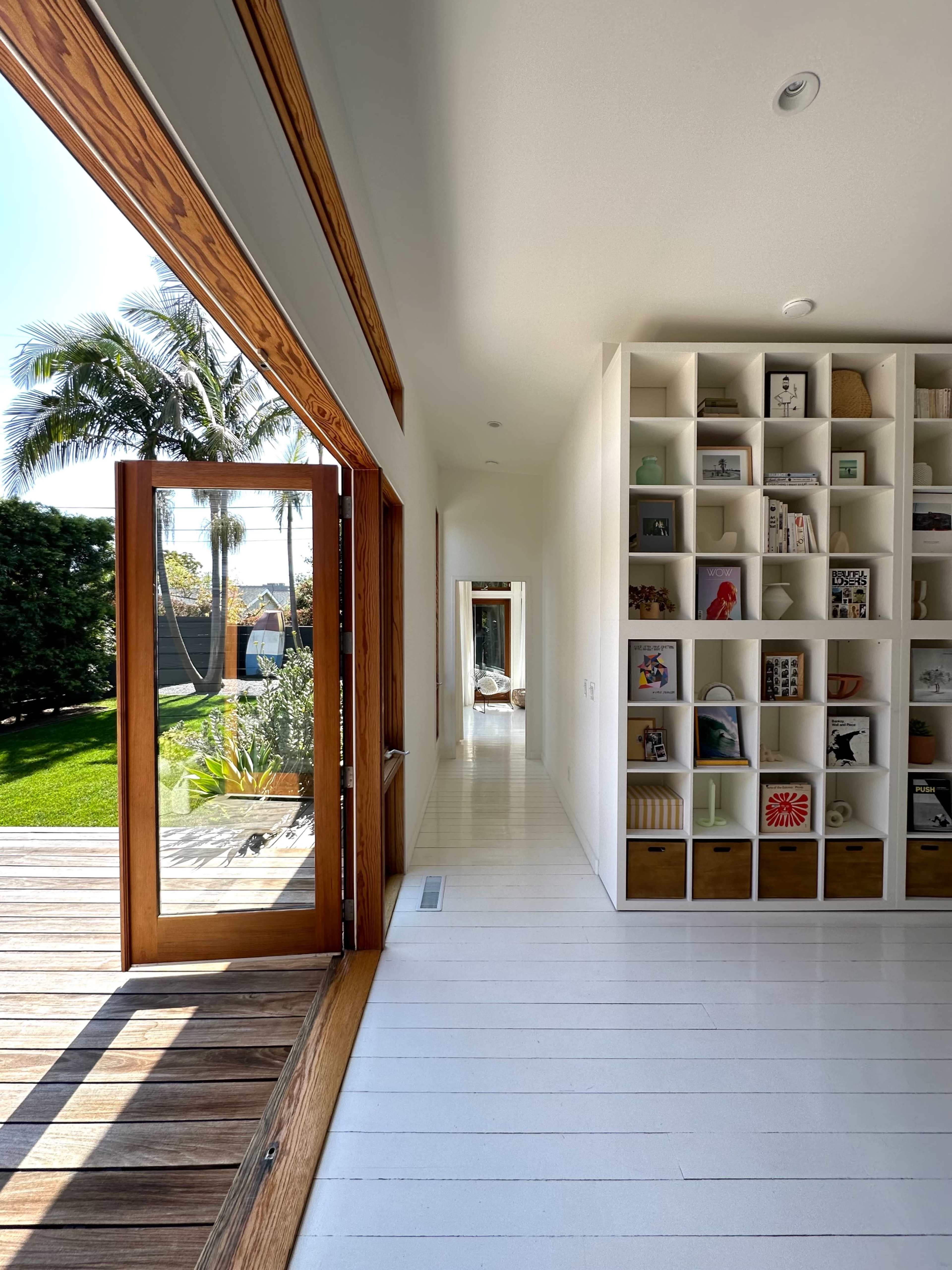 A sunlit hallway features a large open door leading to a wooden deck, with a wall of shelves filled with books and decor on one side.