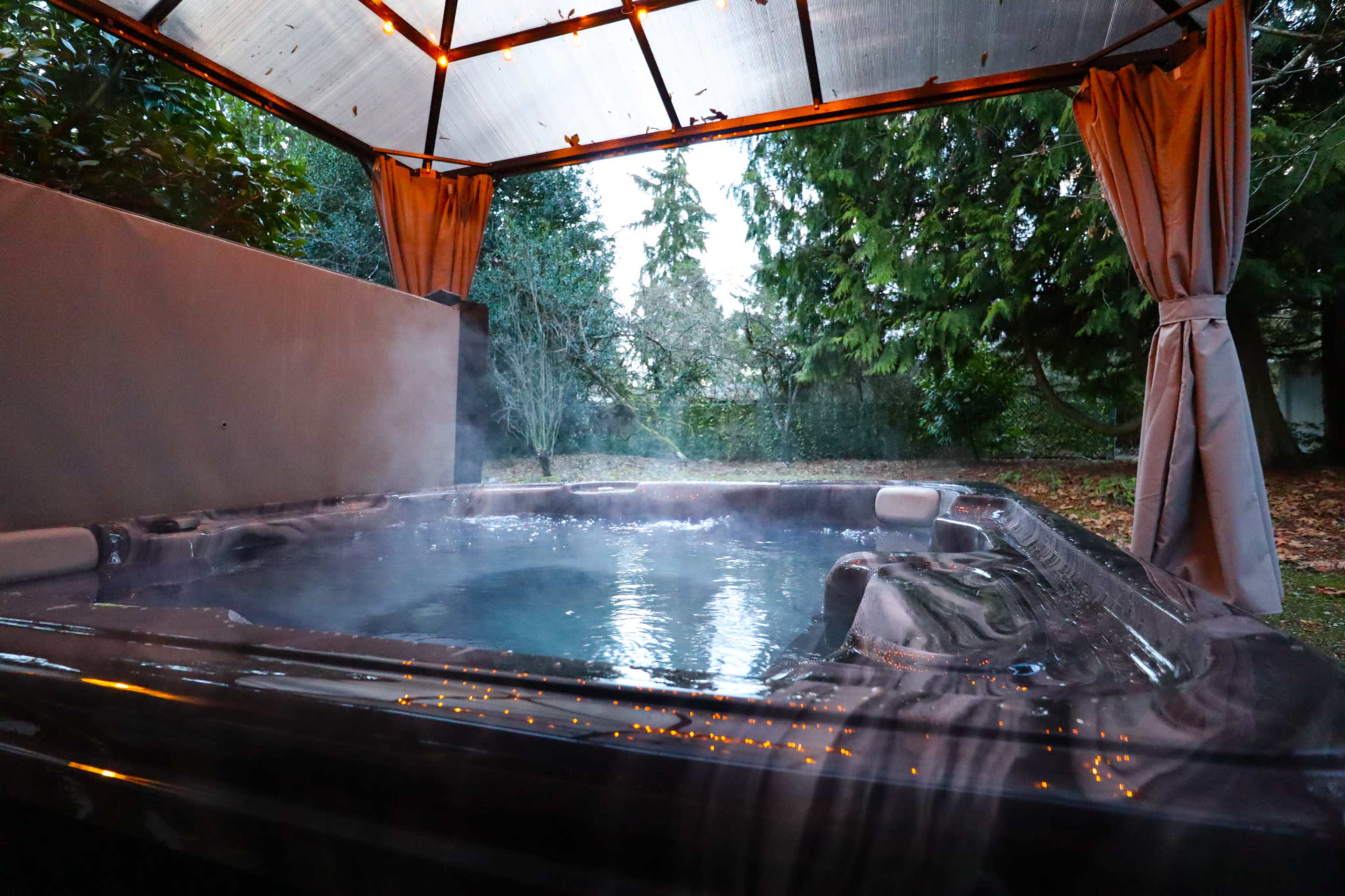 A hot tub with steam rising sits under a covered outdoor area surrounded by greenery.